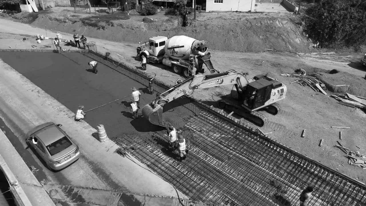 Trabajadores de construcción colocando concreto en una estructura de reja de acero para una carretera, con maquinaria pesada y un camión mezclador de cemento en el fondo.