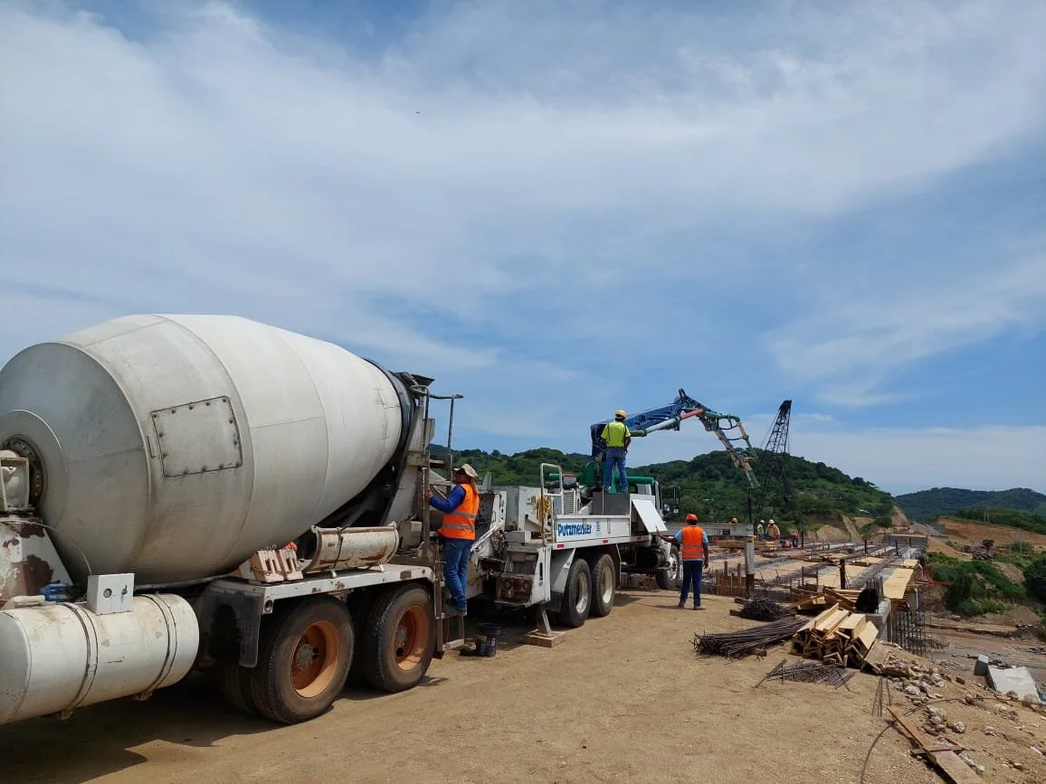Obra de construcción con maquinaria pesada y obreros trabajando en una estructura de concreto en un sitio al aire libre con montañas y cielo despejado en el fondo.