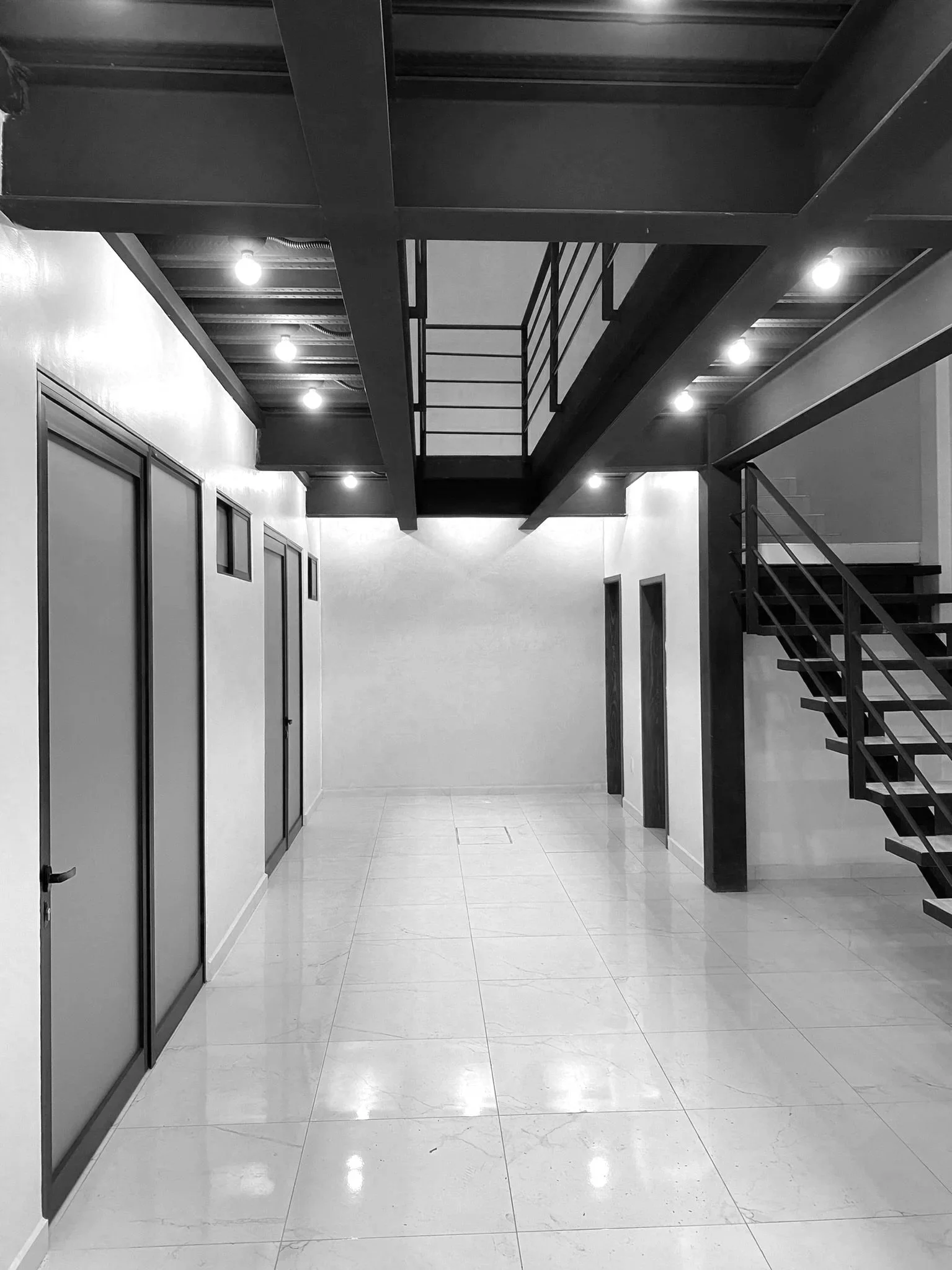 Pasillo interior de edificio con puertas a la izquierda, escalera metálica y estructura en segundo nivel, techos con luces.