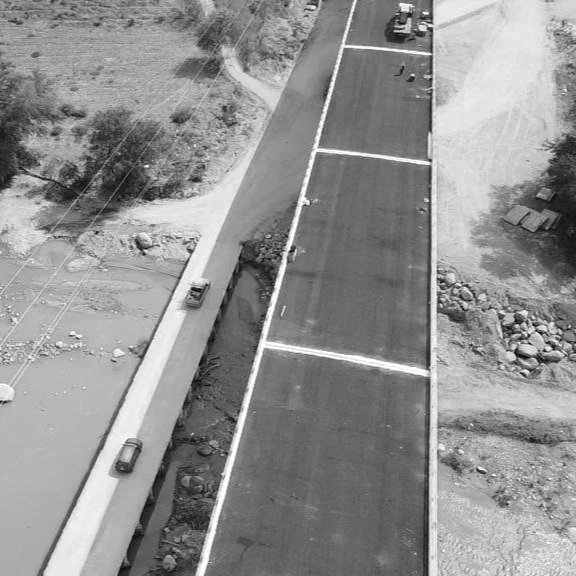 Vista aérea de un puente en construcción con algunos autos en la vía y áreas de tierra y rocas a los lados.