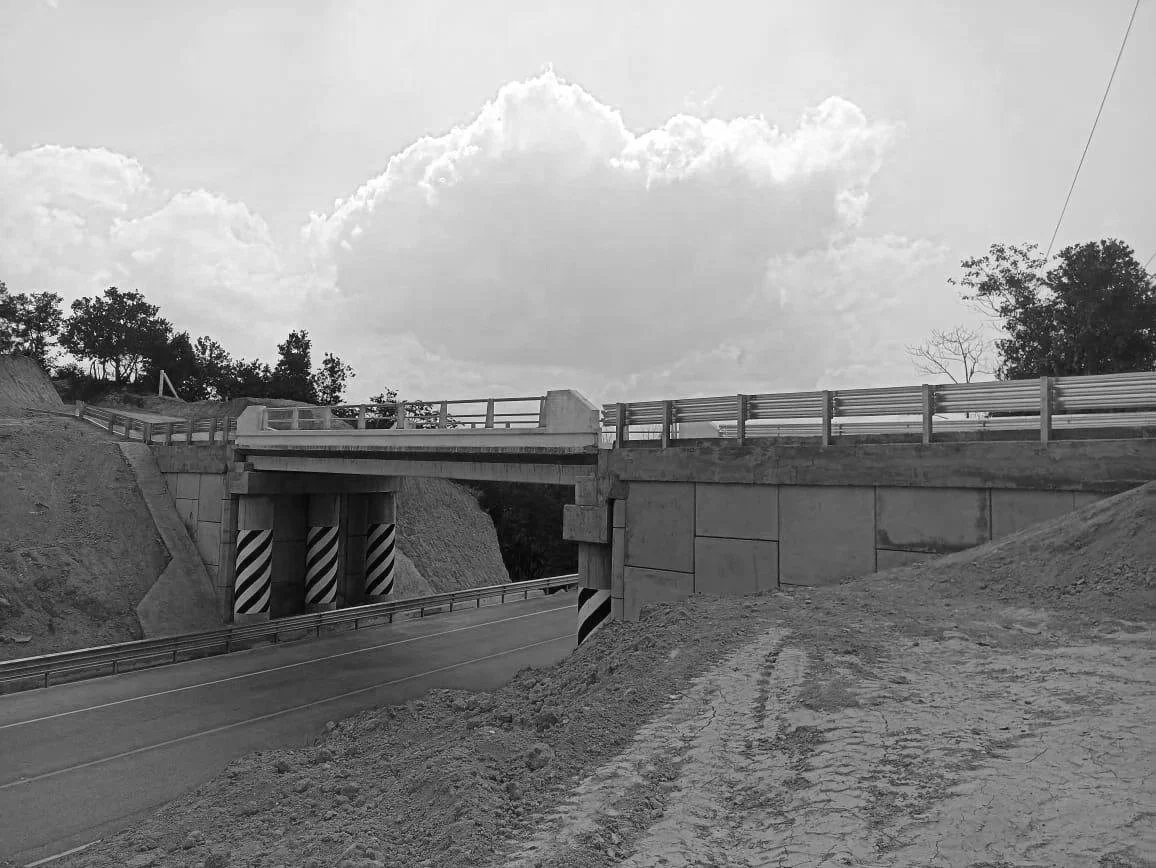 Puente en construcción sobre una carretera en un entorno rural con árboles y cielo nublado.