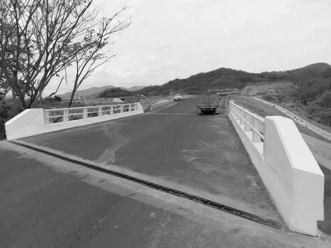 Puente pequeño con barandillas blancas, en una zona rural con montañas y árboles, sin tráfico