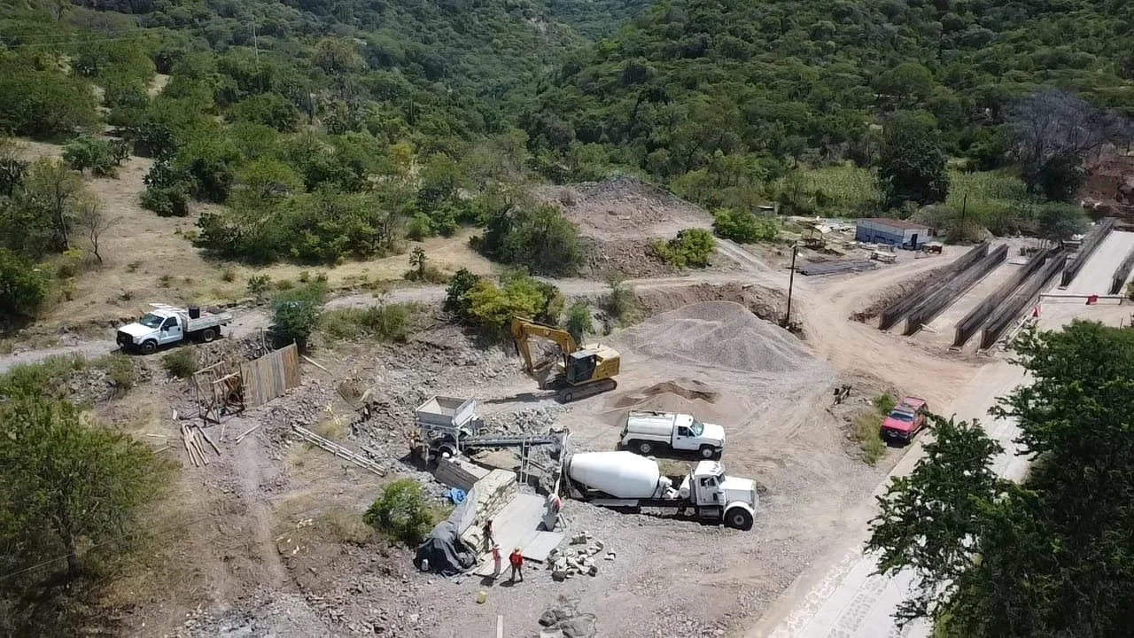Imagen de una obra de construcción en un área rural con maquinaria, camiones y trabajadores, rodeada de árboles y colinas.