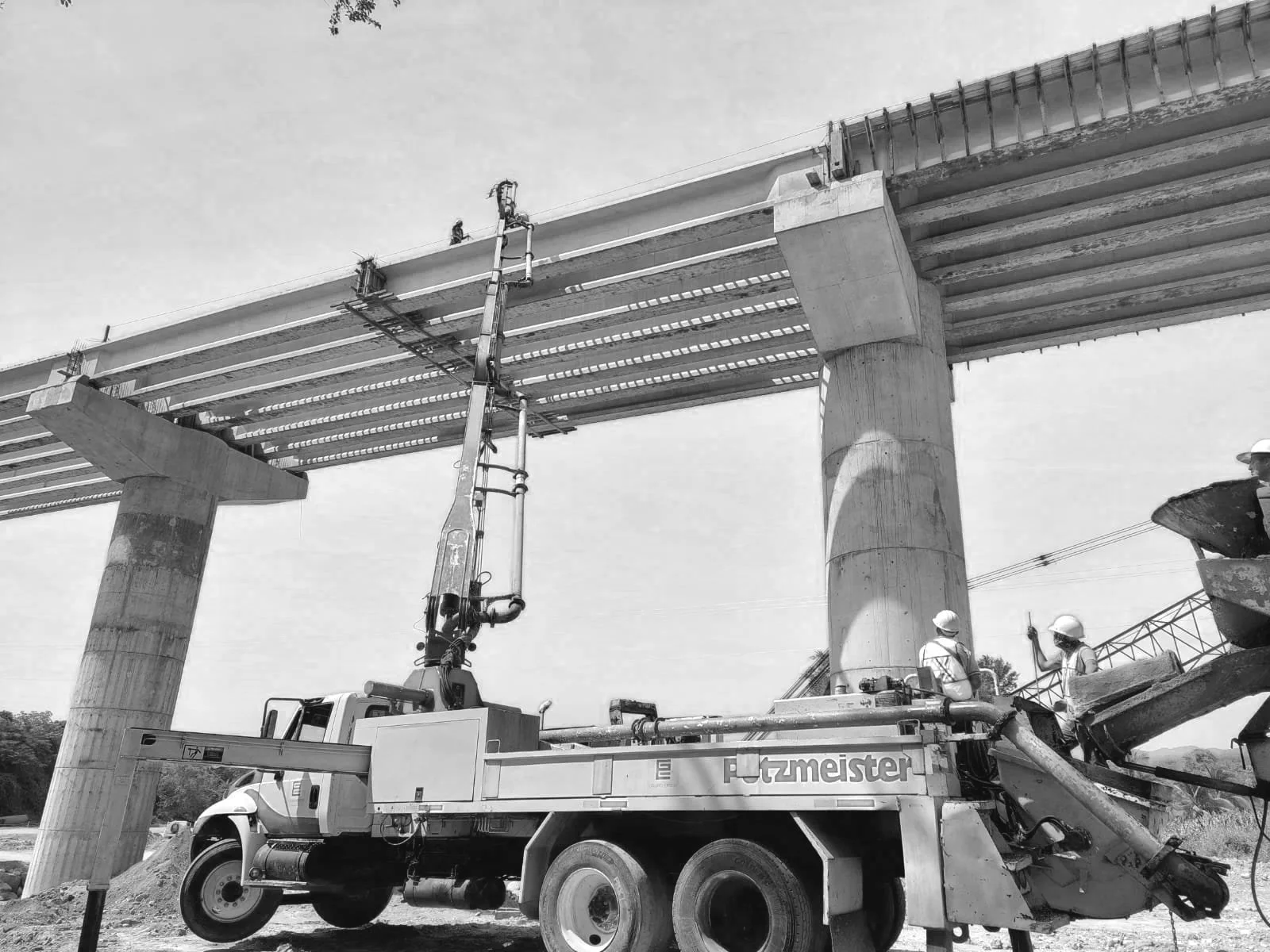 Construcción de un puente con columnas de concreto y estructura de acero, operarios con casco trabajando en el sitio.