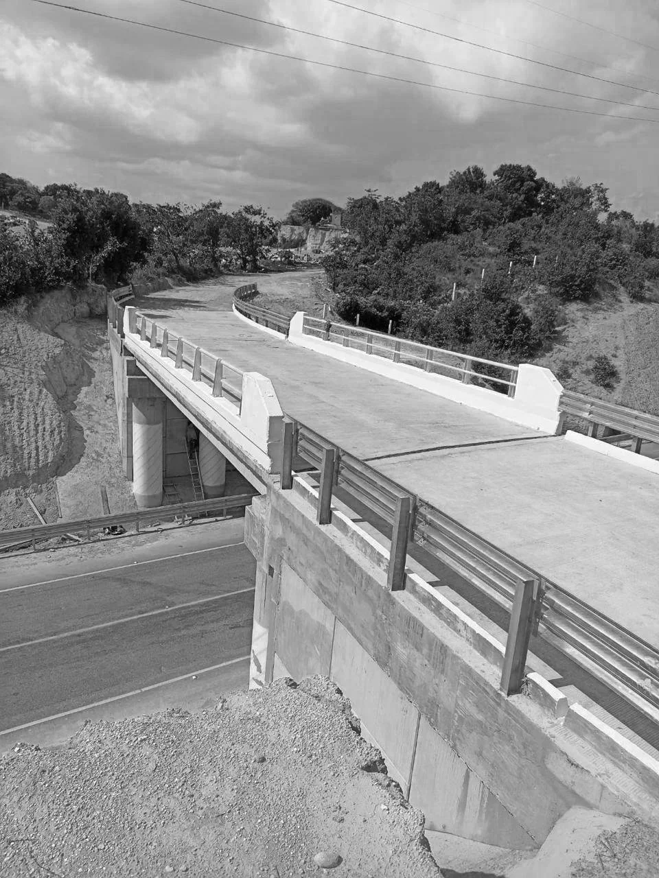 Puente en construcción sobre una autopista en un área con árboles y colinas, con cielo nublado.