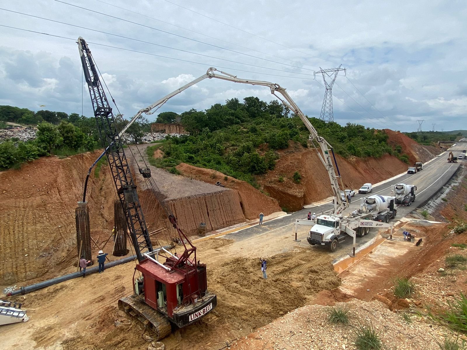 Construcción de un puente vial con maquinaria pesada, incluyendo una grúa y un camión mezclador de cemento, en un área rural con vegetación y una carretera en construcción.