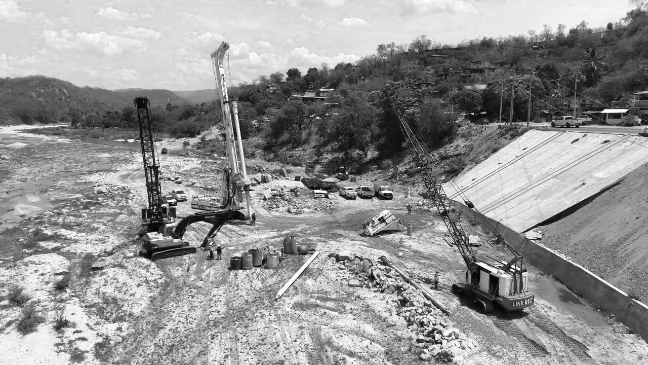 Imagen en blanco y negro de una obra en construcción cerca de un río, con dos grúas y árboles en un paisaje montañoso.