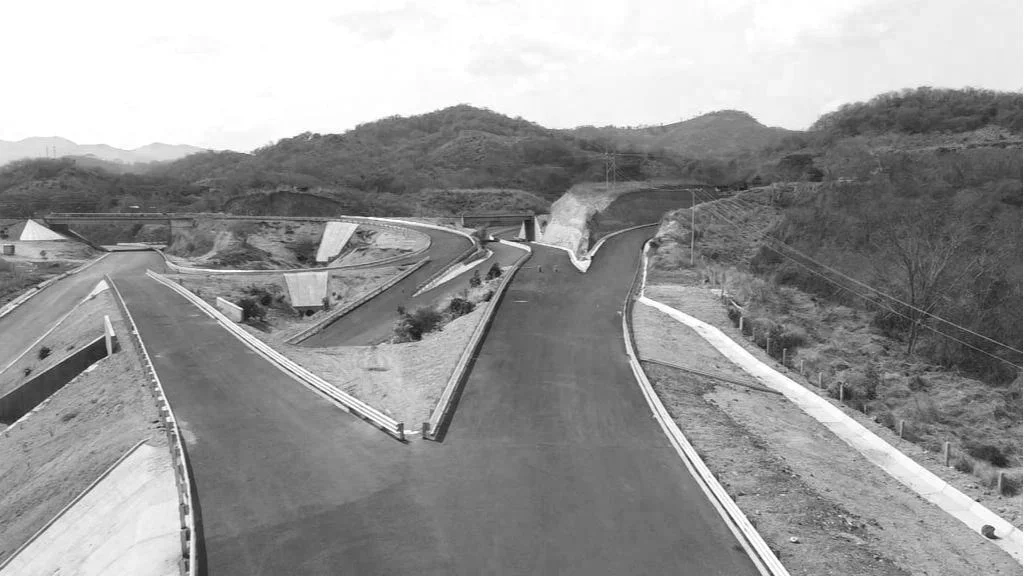 Carretera en construcción en un paisaje montañoso con vegetación escasa en un entorno rural o semiurbano.