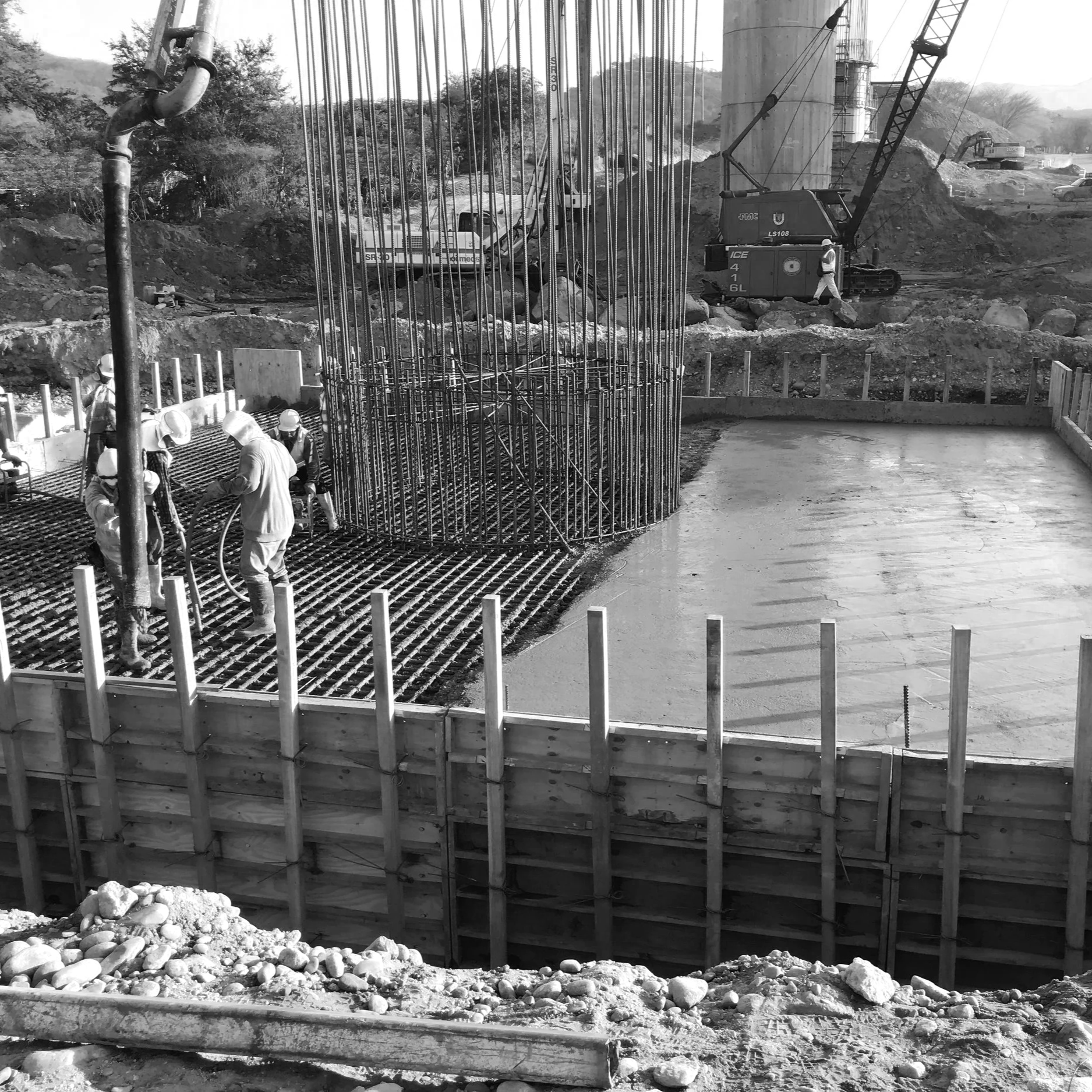 Imagen en blanco y negro de una construcción, con trabajadores en el sitio, algunos vertiendo concreto en una estructura de acero y otros operando maquinaria pesada en el fondo.
