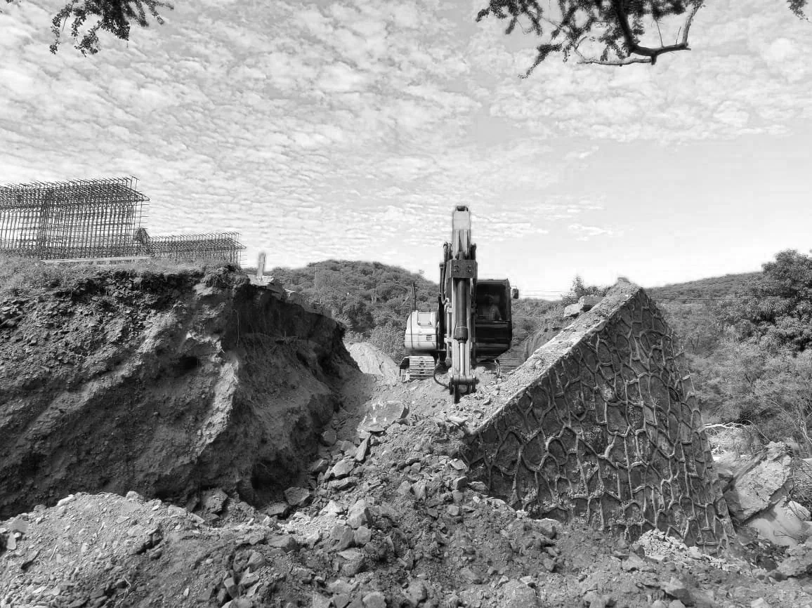 Excavadora trabajando en un proyecto de construcción de caminos, con estructuras de acero en el fondo y un cielo nublado.