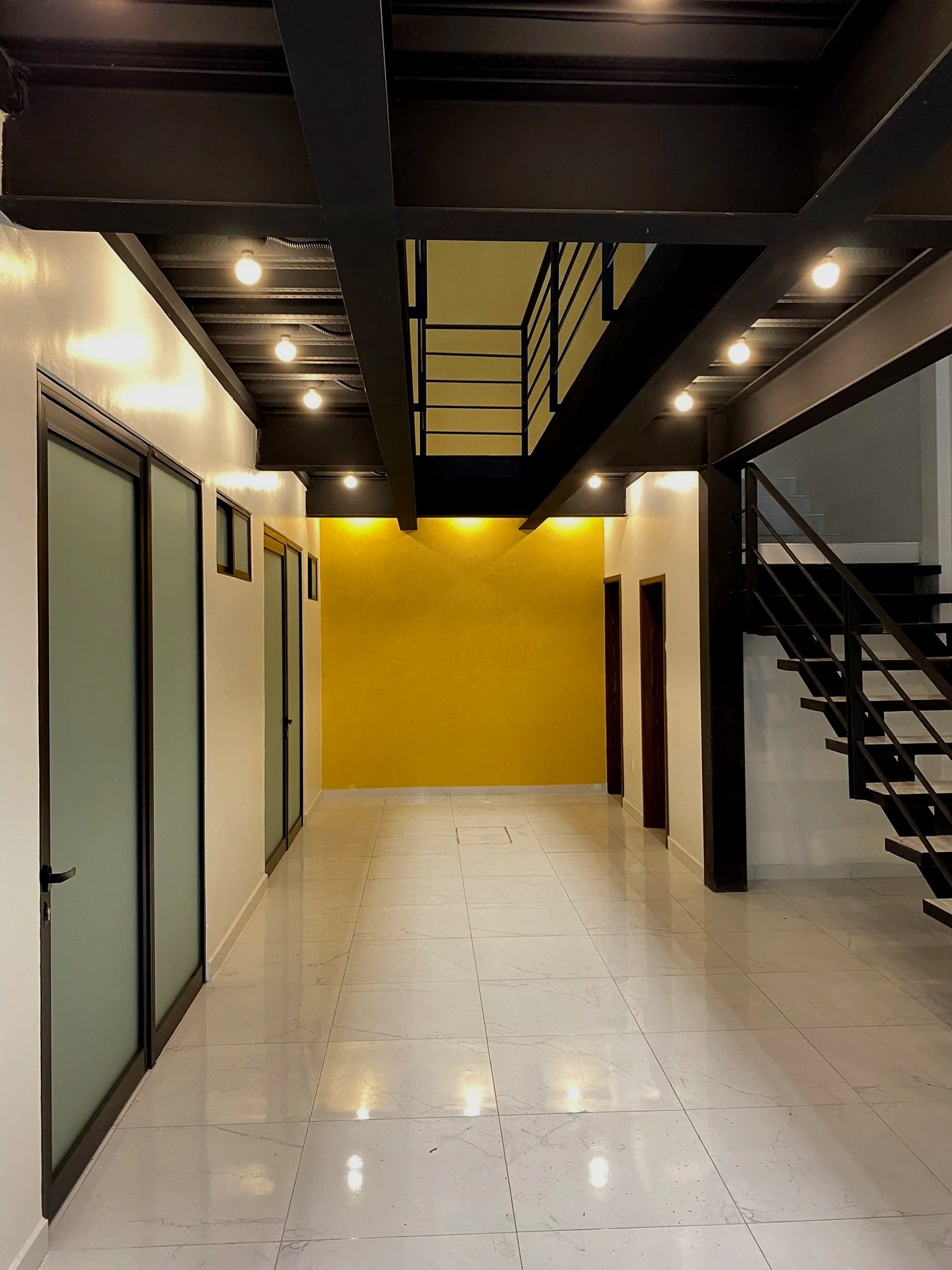 Interior moderno con paredes blancas, una pared amarilla, suelo de baldosas crema, puertas de vidrio con marco negro, escaleras negras con barandilla y luces empotradas en el techo de estructura negra.
