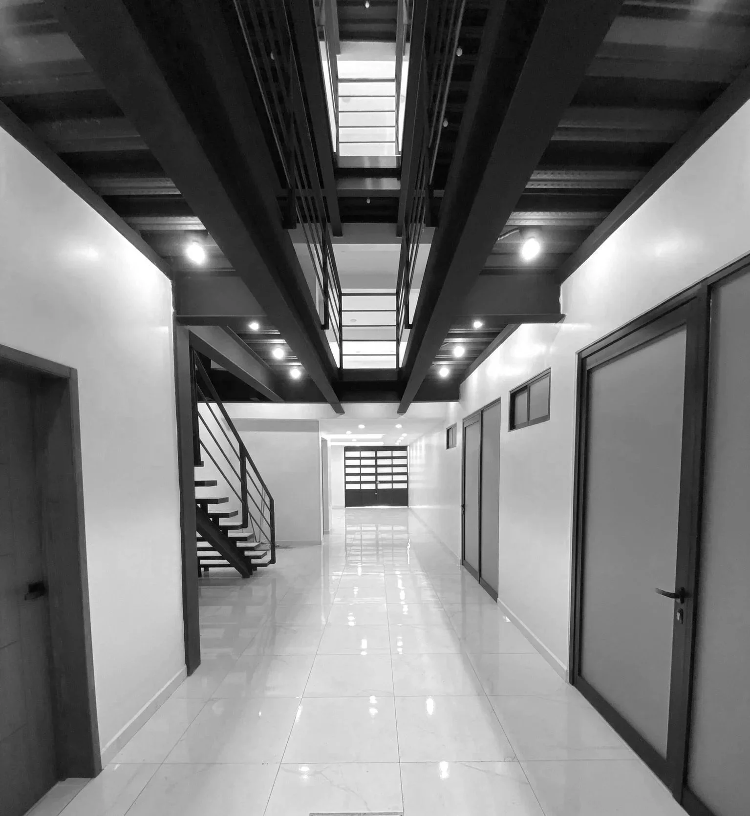 Interior de un edificio con pasillo amplio, puertas de madera y ventanas altas, escalera y estructura de metal en el segundo piso, iluminación moderna.