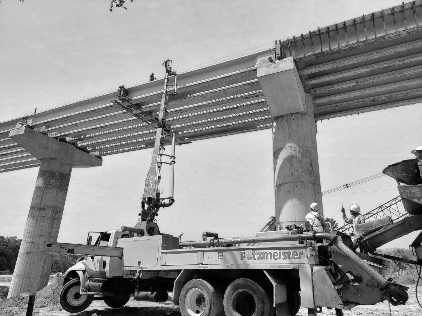 Construcción de un puente con pilotes de concreto y estructura metálica, con trabajadores en casco de seguridad y maquinaria en un sitio de obra.