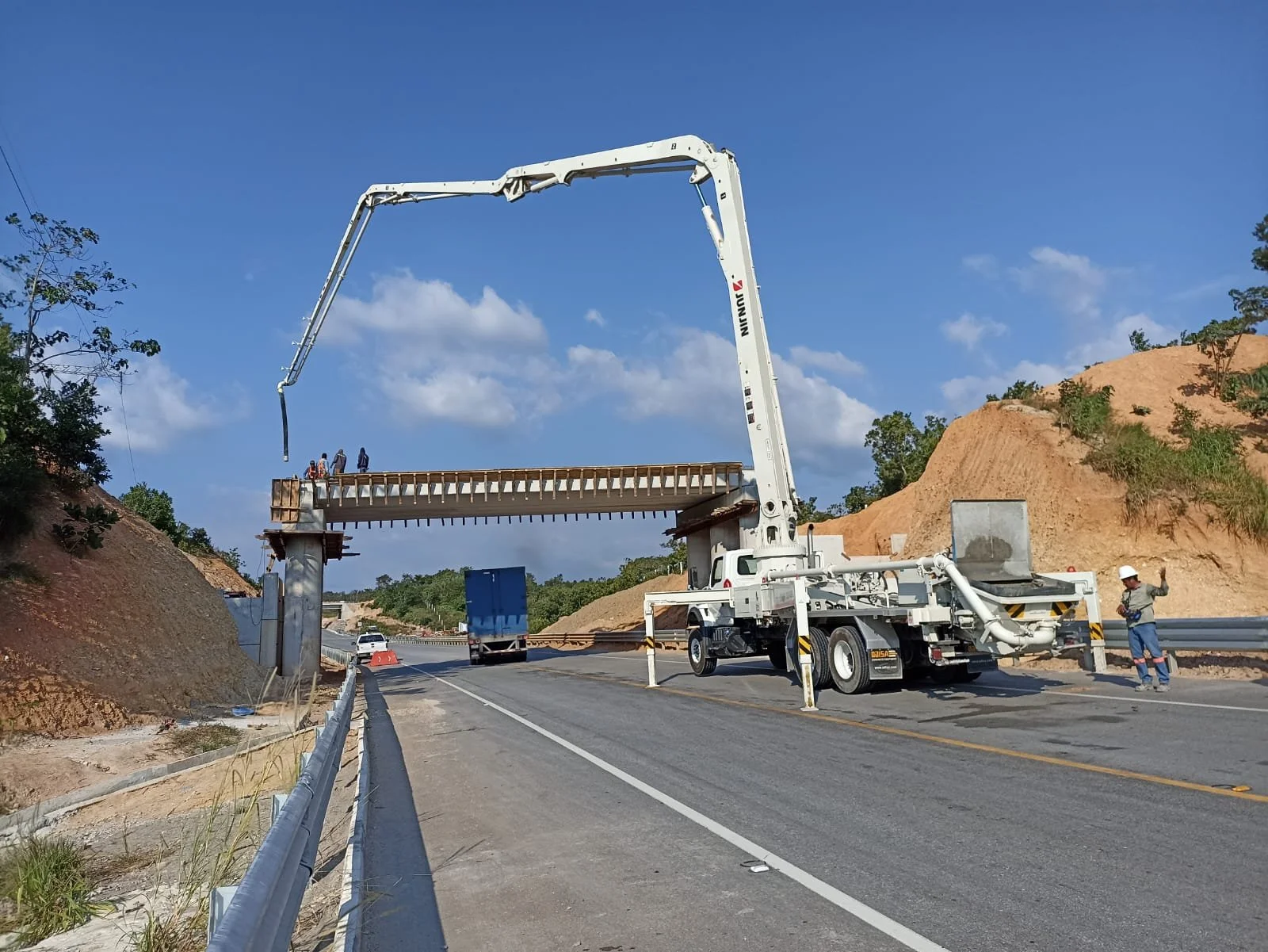 Personal de construcción y maquinaria trabajando en la estructura de un puente en una autopista, con colinas y árboles en el fondo en un día soleado.
