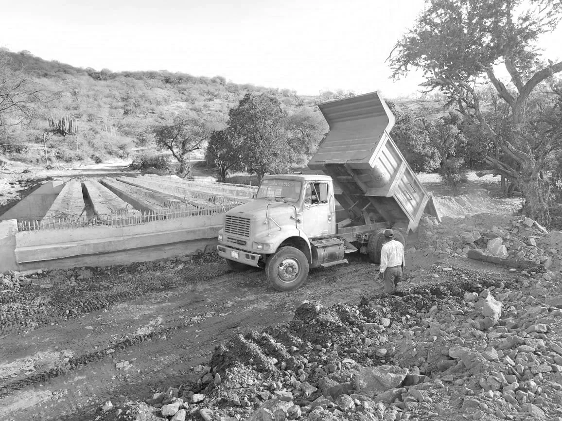 Camión volcado en un camino de tierra, con un conductor y un acompañante en un área rural con árboles y colinas en el fondo.