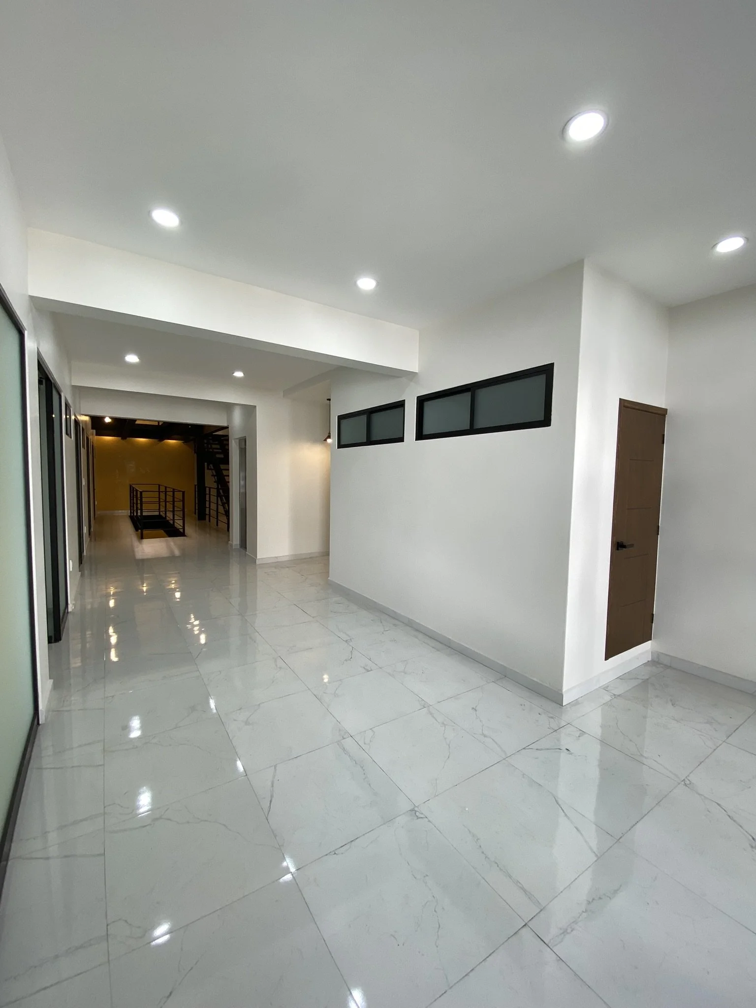 Interior de una casa moderna con paredes blancas, suelo de mármol brillante, ventanas pequeñas y luces en el techo. Se observa un pasillo y una escalera en el fondo.