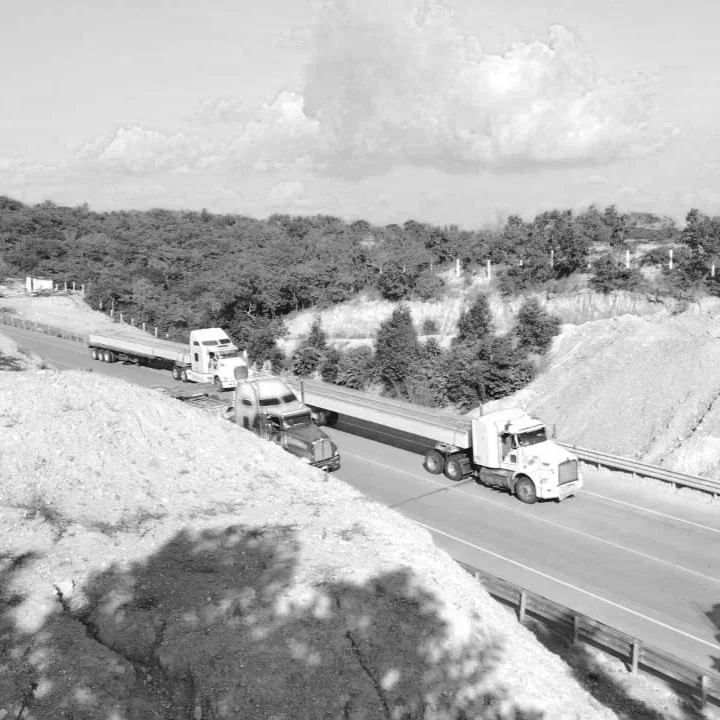 Camiones y autos en una carretera con paisaje montañoso y árboles, en un día soleado con nubes en el cielo.
