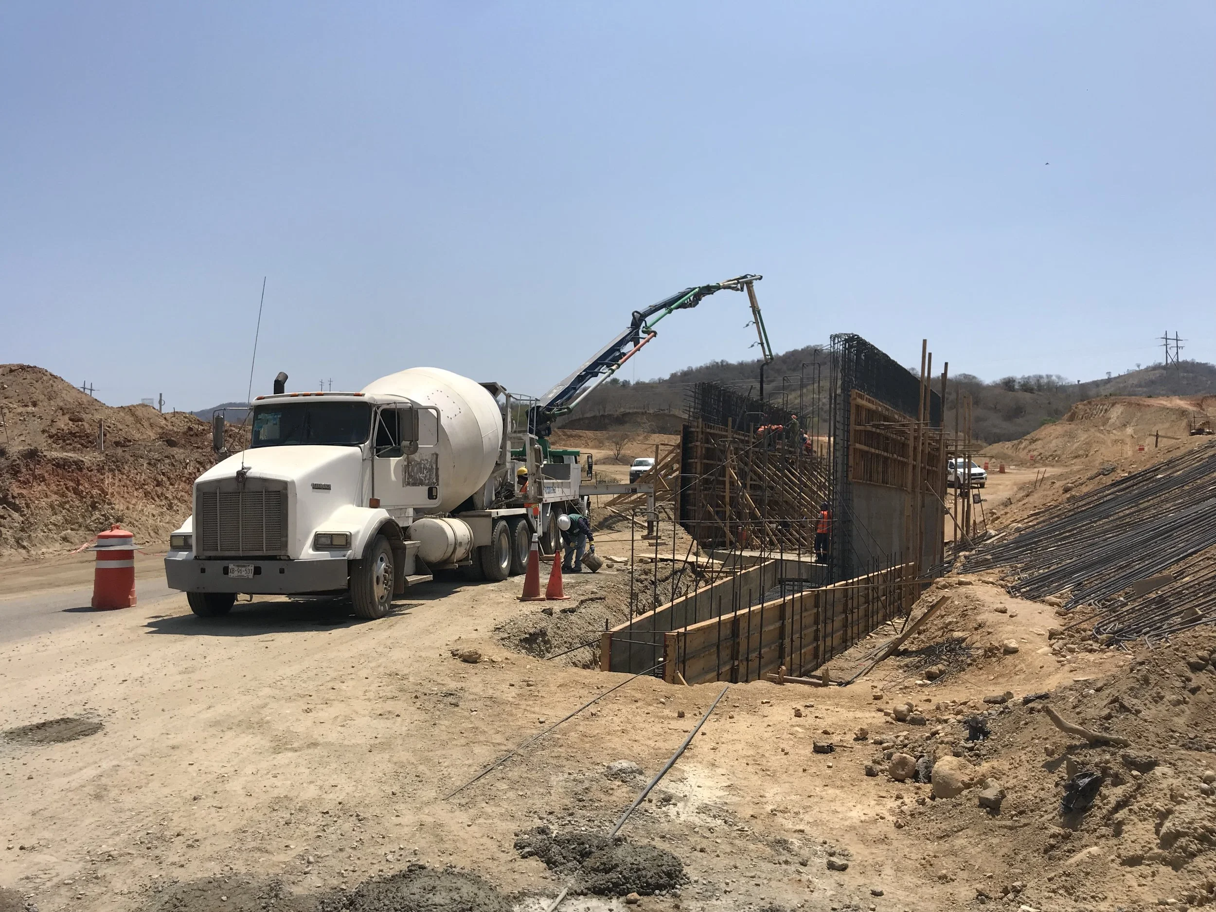 Camión mezclador de cemento vertiendo concreto en una estructura en construcción, con trabajadores en el sitio y un cielo despejado en un área de terracería.