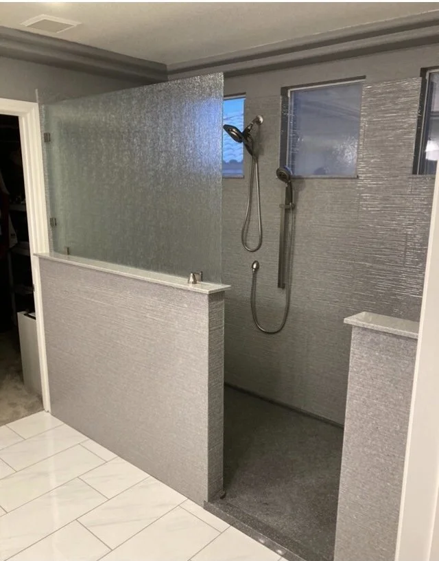 Modern barrier-free walk-in shower with half wall glass enclosure, dual shower heads, and textured wall surround in custom bathroom remodel