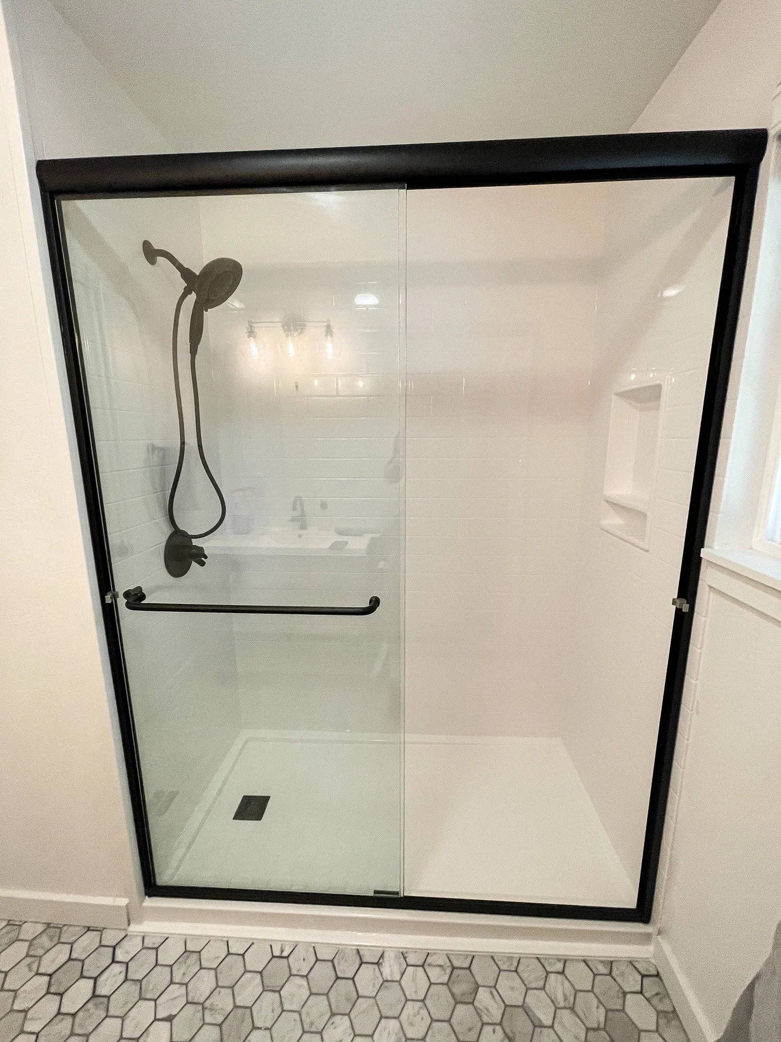 Modern sliding glass shower door with matte black frame, white tile walls, and hexagon tile flooring in a remodeled bathroom.