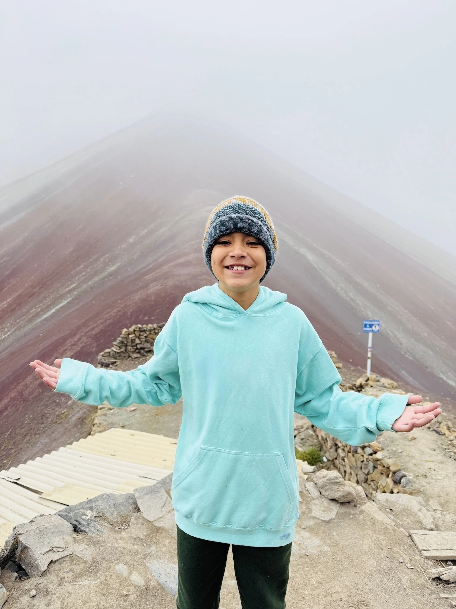 Boy standing at Vinicunca (Rainbow Mountain) in the Peruvian Andes with colorful mountain slopes behind
