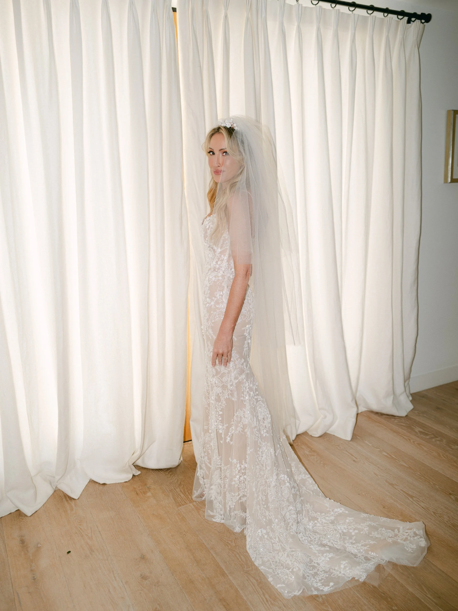 A bride in a white lace wedding dress with a long train and a veil, standing indoors in front of cream-colored curtains.
