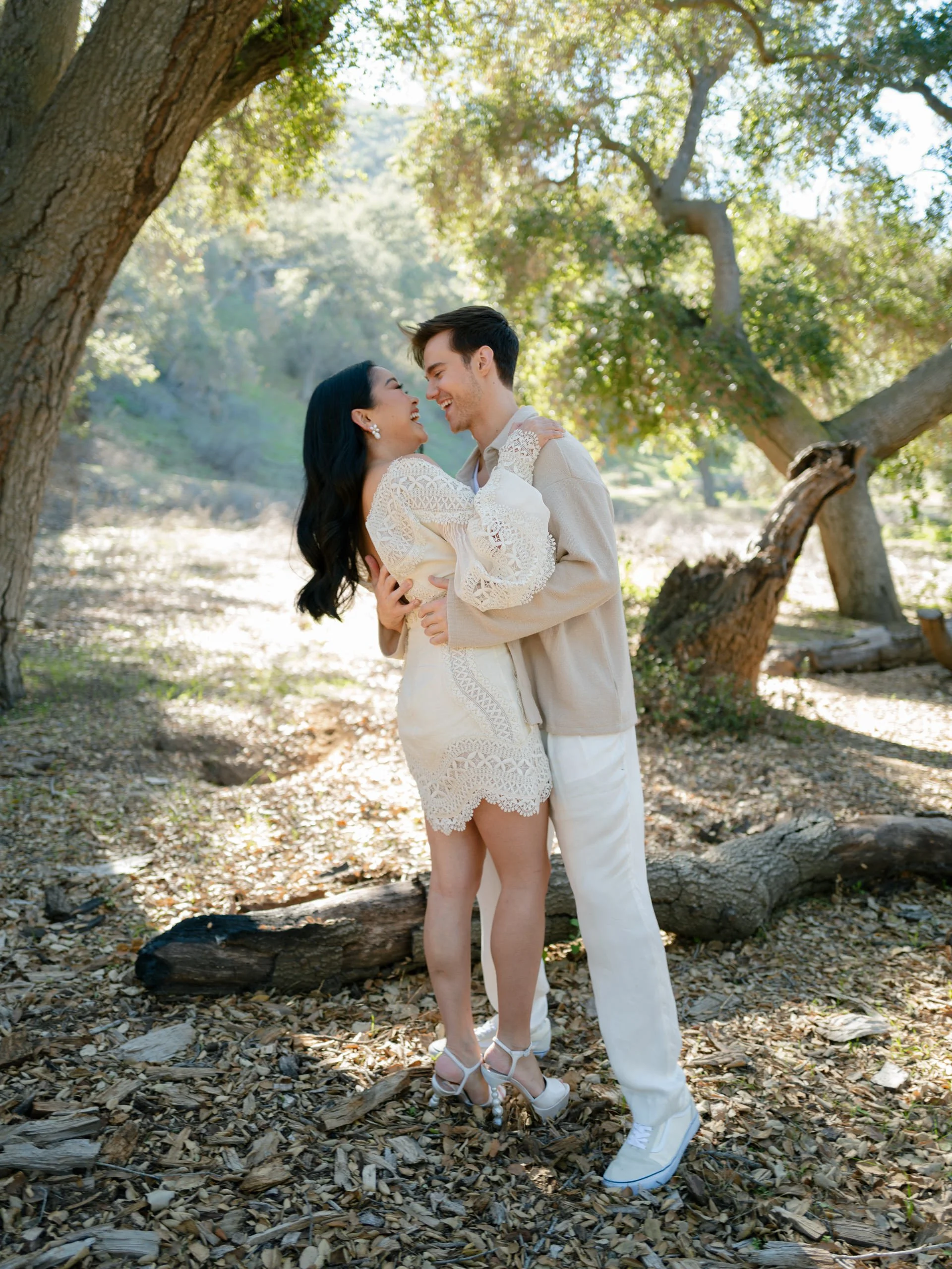 A happy couple is embracing and smiling at each other outdoors under large trees, with sunlight shining through the leaves in a wooded area.