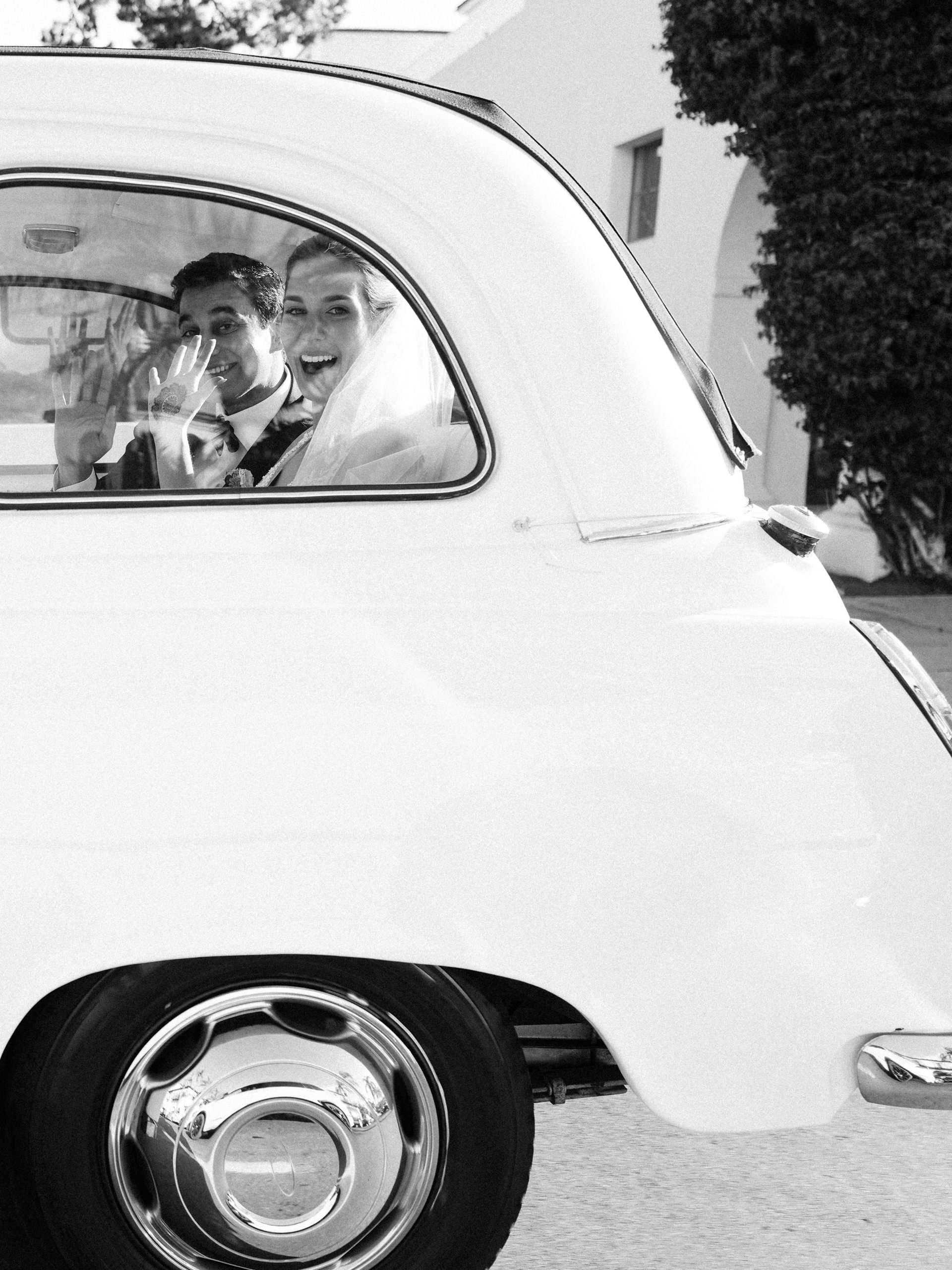 Black and white photo of a smiling bride and groom inside a vintage car, waving through the window.