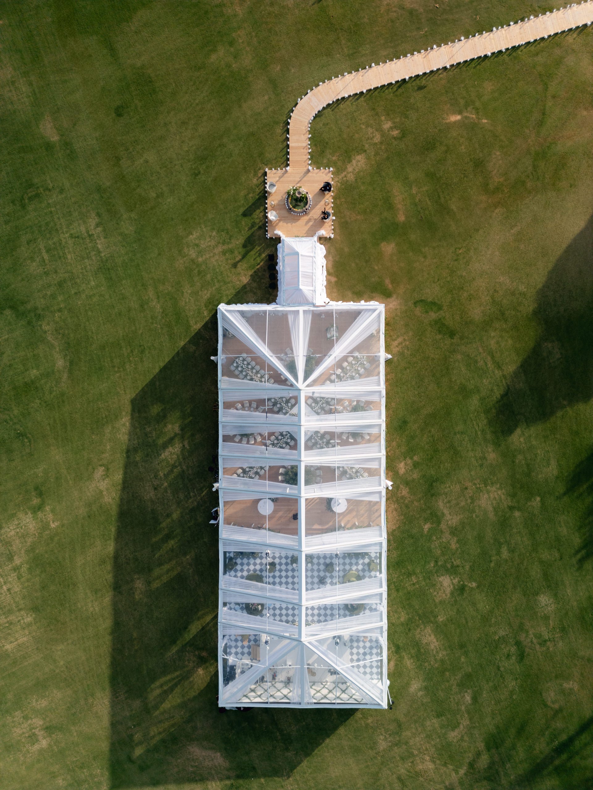 Aerial view of a white outdoor event tent with glass windows on a grassy lawn, connected to a small wooden deck with a pathway leading to a larger deck with tables and chairs.