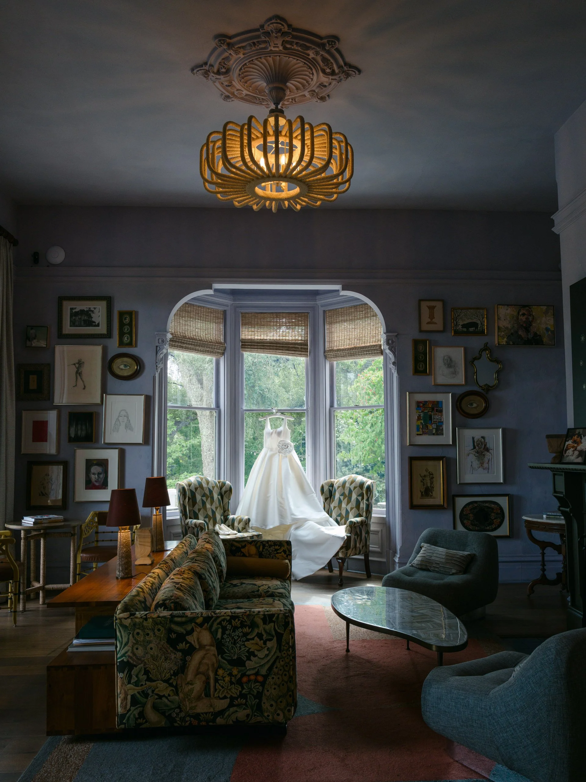 A wedding dress displayed on a window ledge in a cozy living room with framed art, vintage furniture, and a patterned sofa.