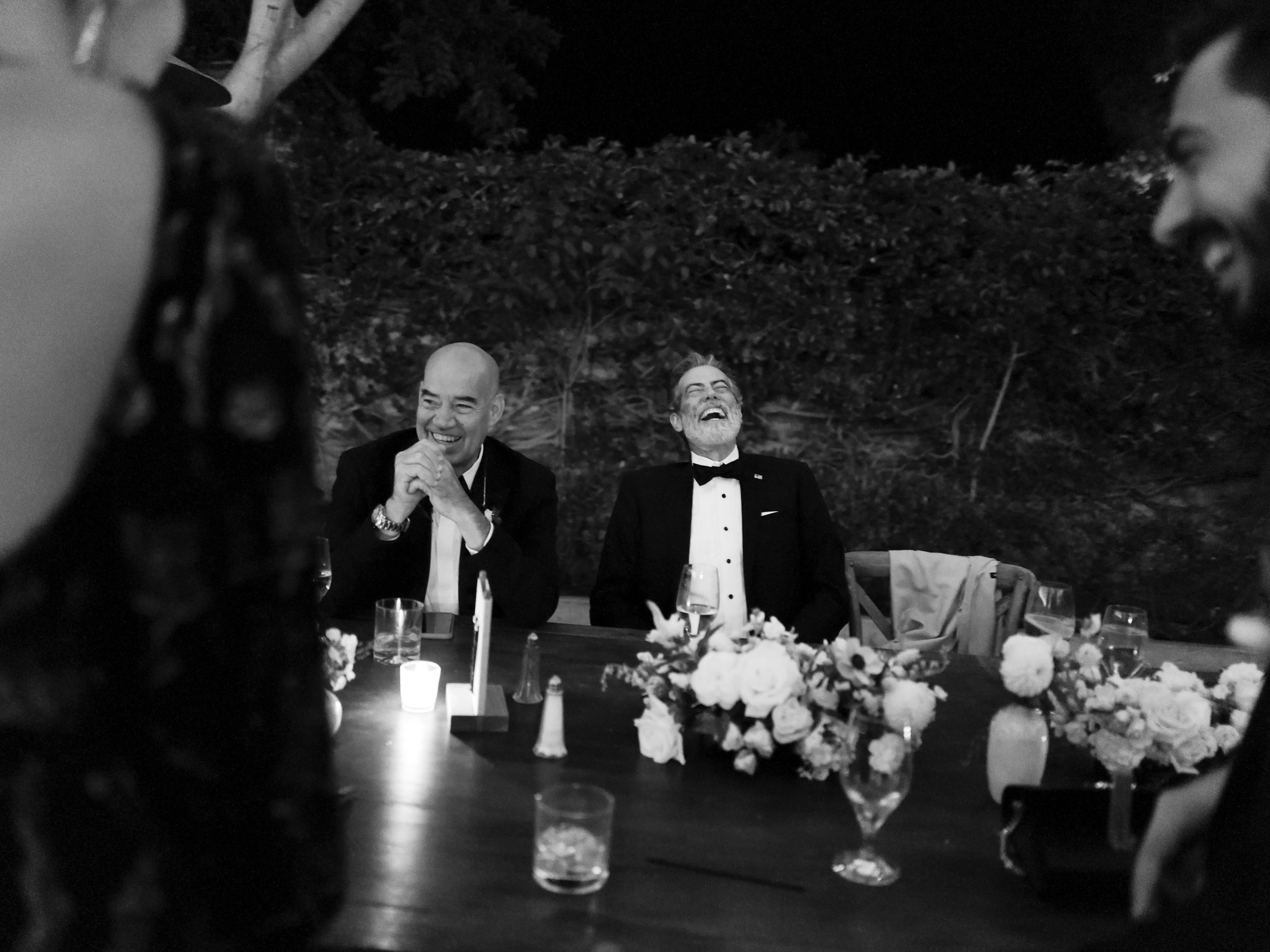 Men dressed in tuxedos at a dinner table, laughing and smiling during an outdoor event at night, with floral arrangements and drinks.
