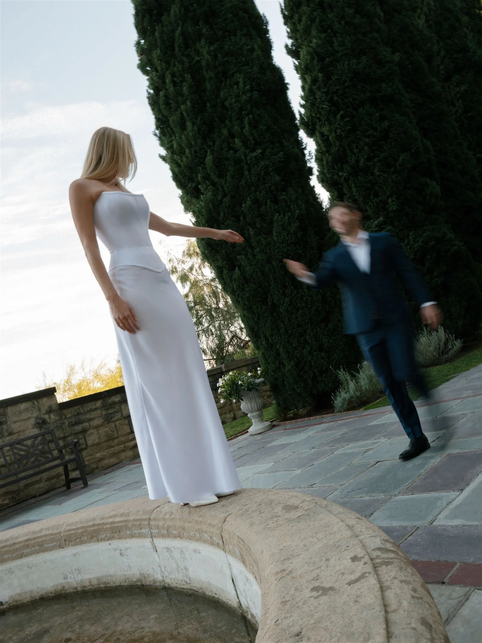 A woman in a white dress standing on a stone ledge and a man in a dark suit running near her outside in a garden with tall trees and a stone wall.