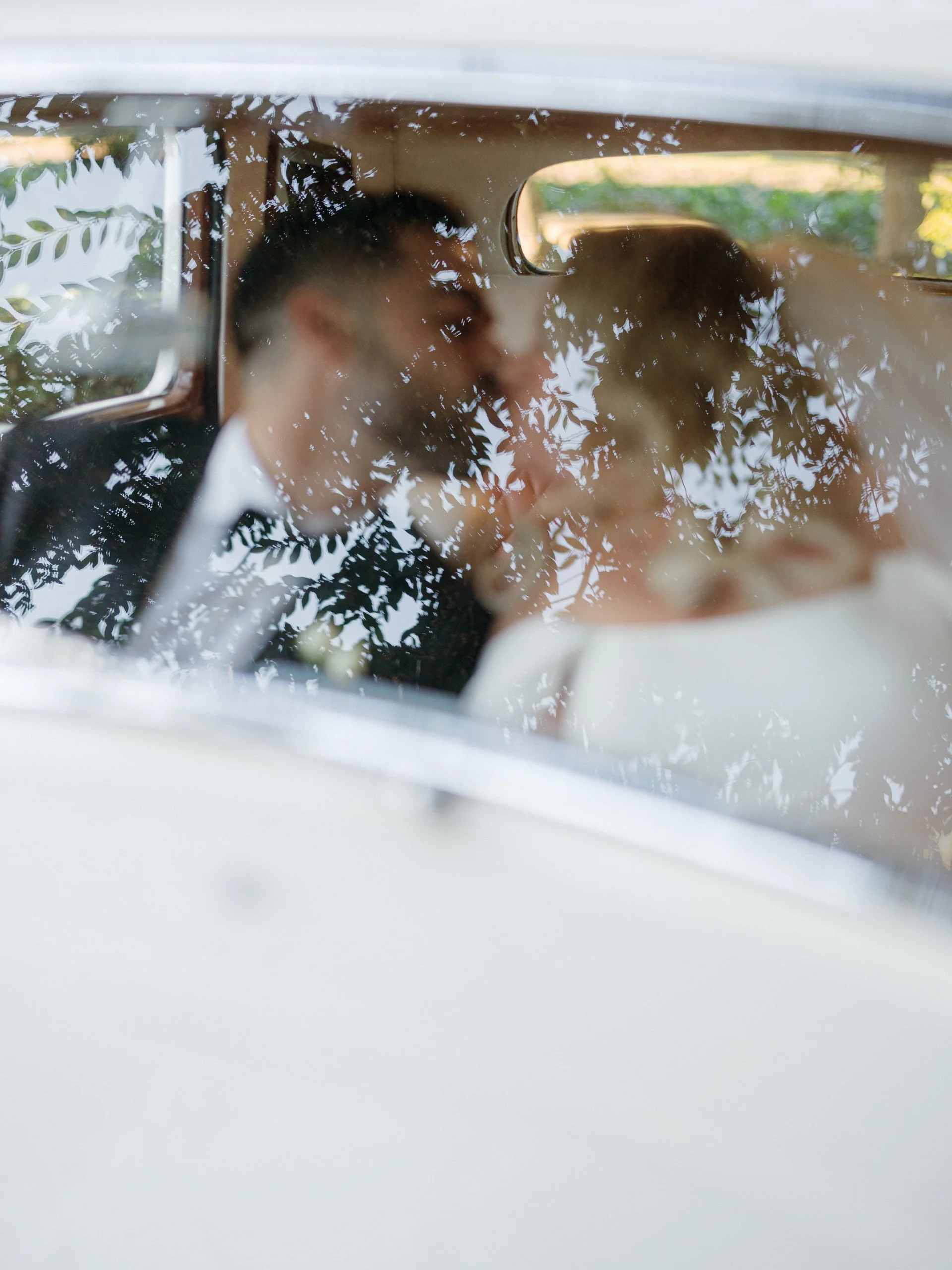 A bride and groom sharing a kiss inside a car, with reflections of tree branches on the window.