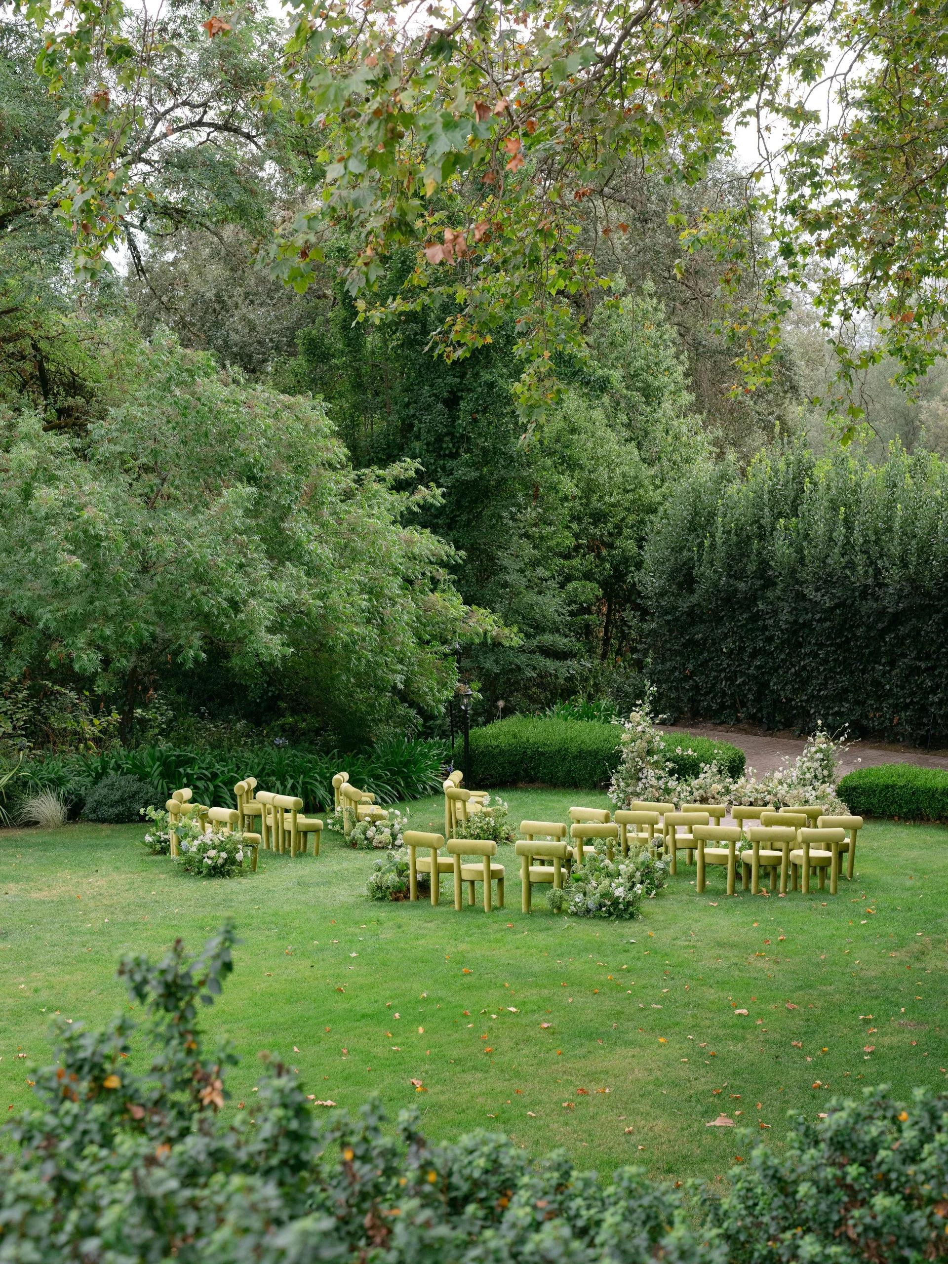 Outdoor garden setup with yellow chairs and floral arrangements for a wedding or event, surrounded by lush green trees and bushes.