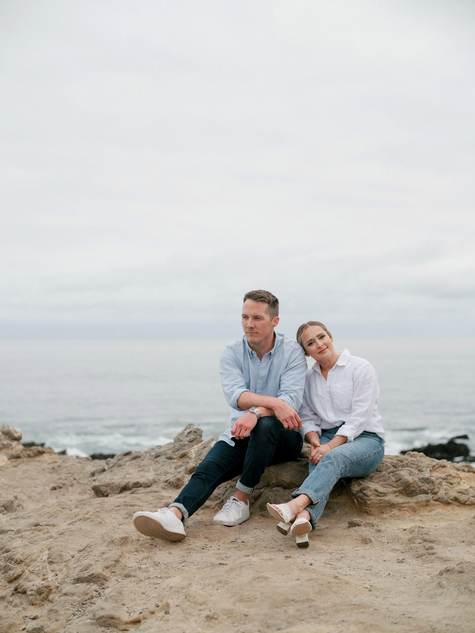 A young couple sitting on rocks by the beach with an overcast sky.