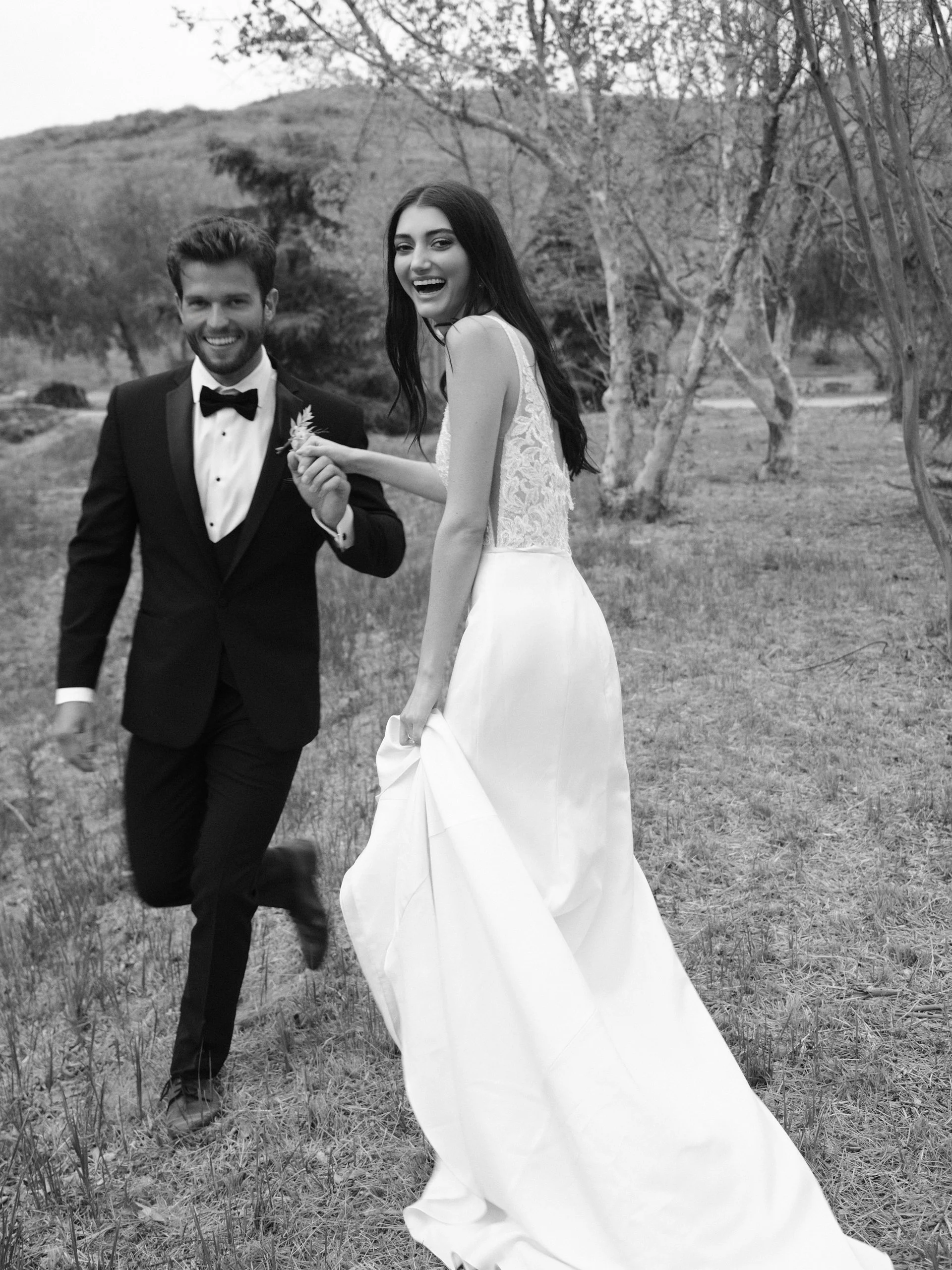 A couple in wedding attire holding hands and smiling outdoors, with trees and grass in the background.