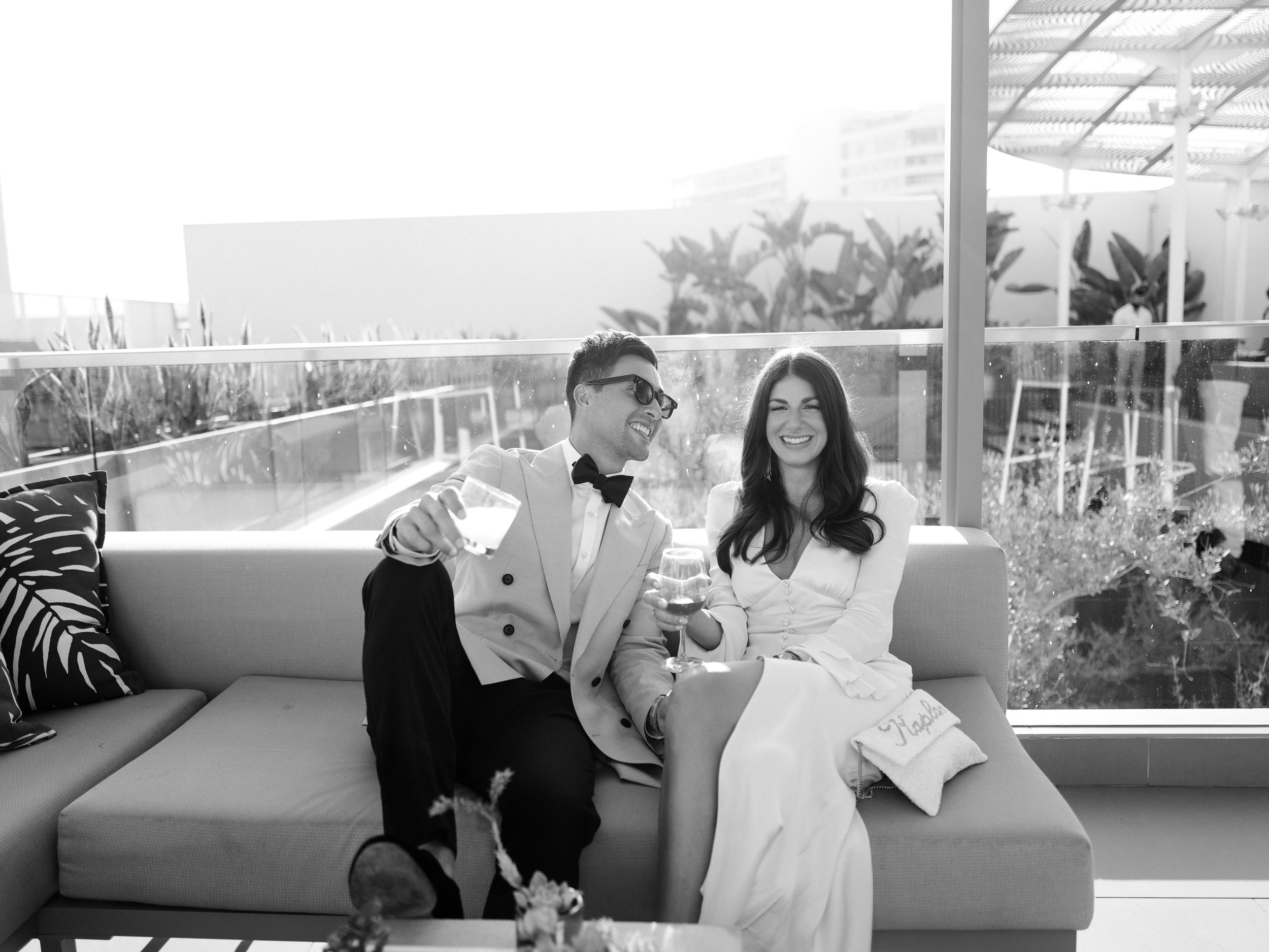 A couple in formal attire sitting on a couch, smiling and enjoying drinks, with a glass barrier and a cityscape in the background.