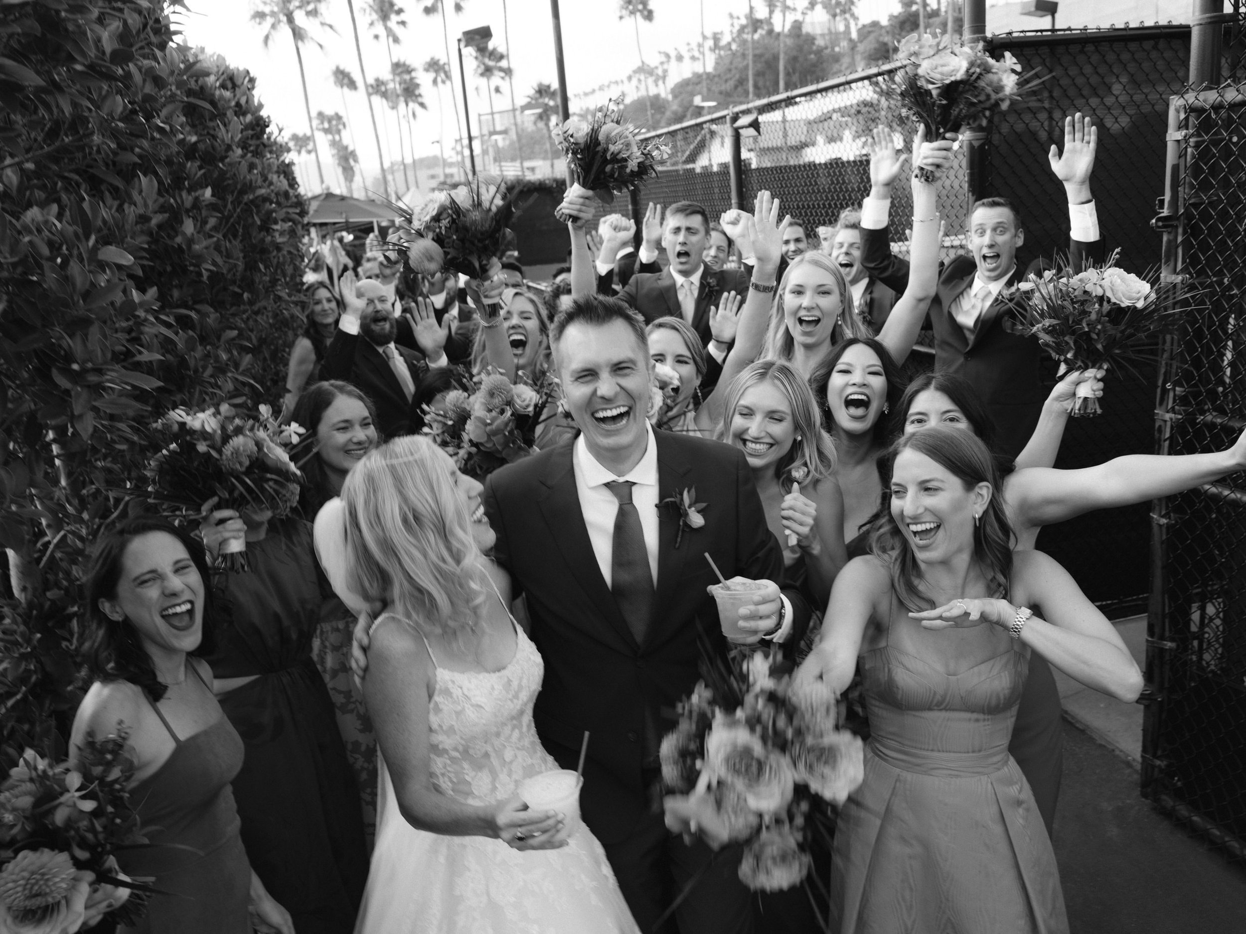 A joyful wedding celebration with a group of smiling people, including the bride and groom, holding flowers and drinks, with some raising their hands, outdoors with palm trees in the background.