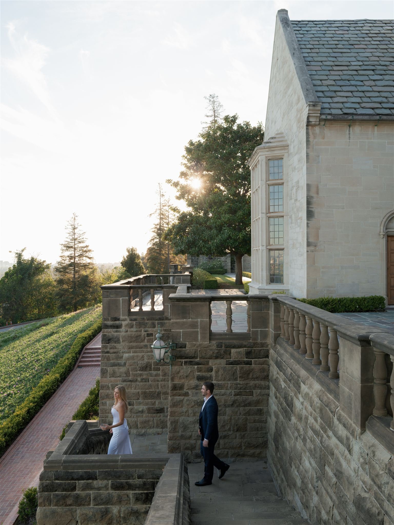 A bride and groom walking down the stairs outside a stone mansion with a garden and trees in the background, sunlight shining through the trees.