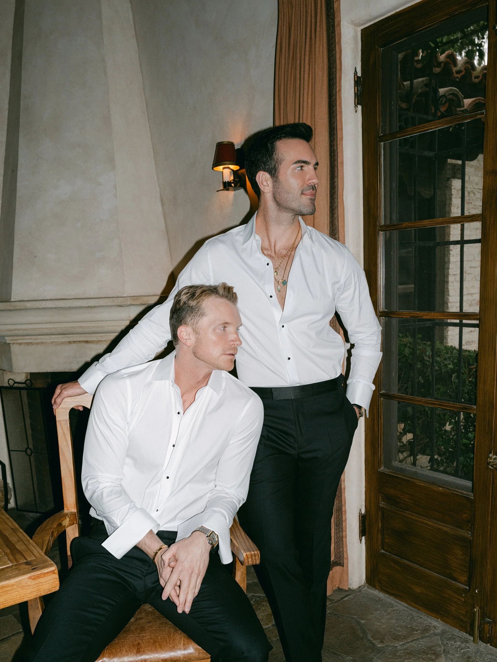 Two men in white shirts and black pants in a stylish, dimly lit room near a window. One man is seated on a wooden chair, and the other is standing nearby, both looking to the right.