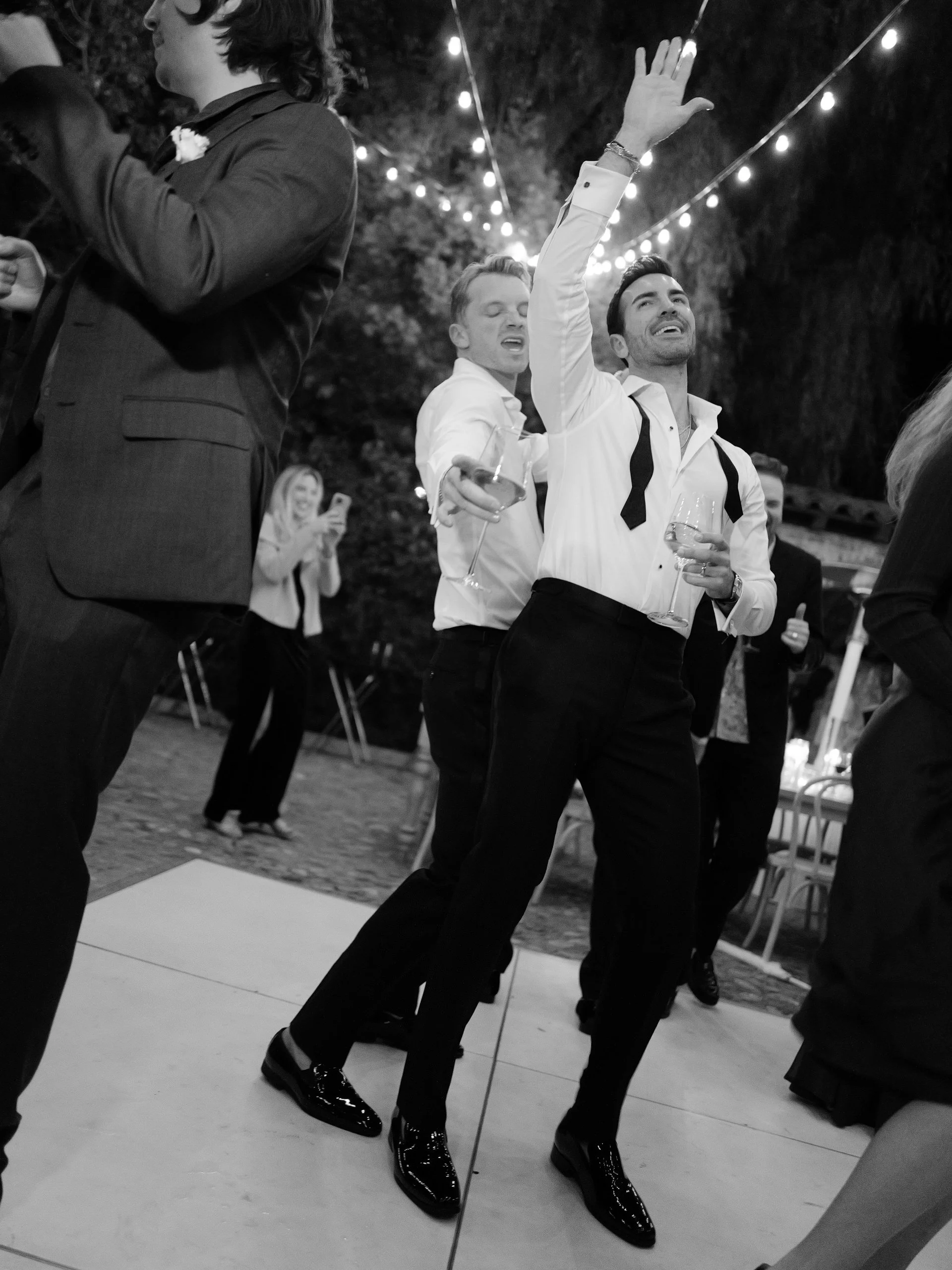 People dancing at an outdoor nighttime event with string lights overhead. Two men are in focus, one with arm raised and the other smiling, holding wine glasses.