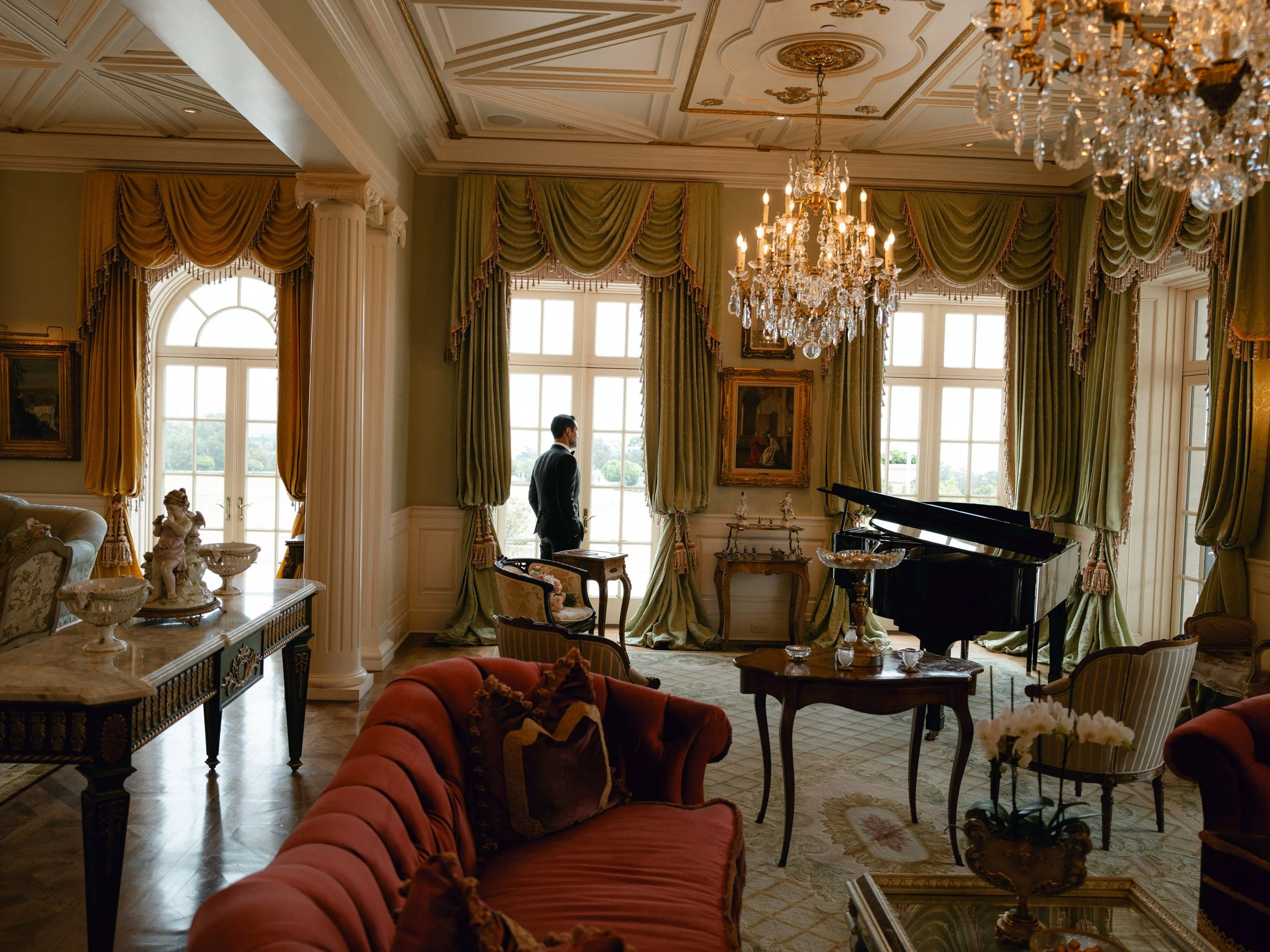 A man stands near large windows in an elegant, ornate room with luxurious furniture, chandeliers, and gold-trimmed curtains.