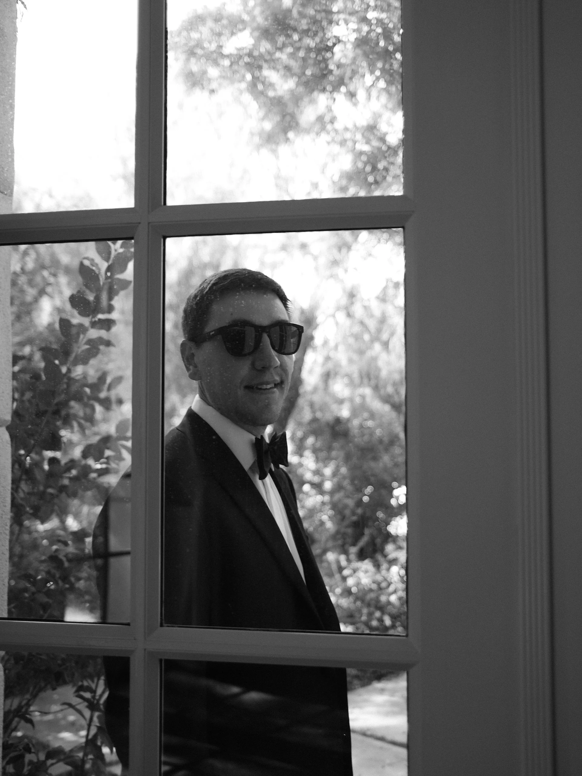A man dressed in a tuxedo with a bow tie and sunglasses is seen through a window with divided panes, with trees and foliage in the background.