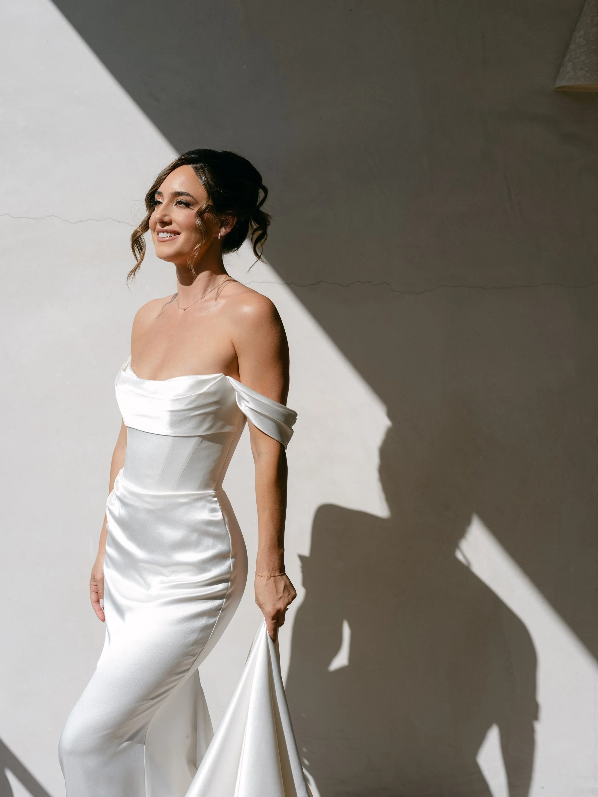 A woman in a white satin off-the-shoulder dress standing against a wall with sunlight creating a shadow of her outline.
