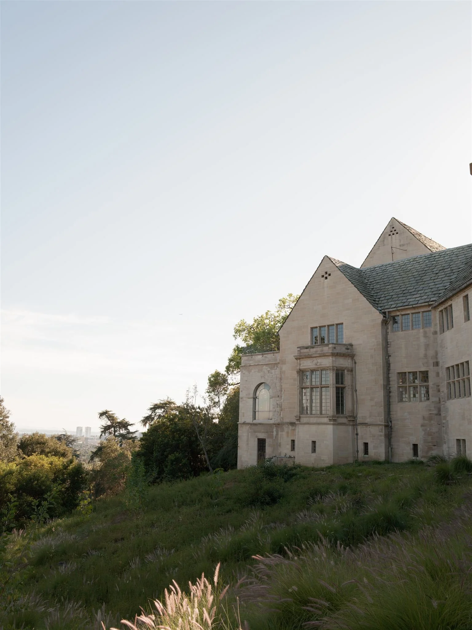 A historic stone mansion on a hillside with a lush garden, trees, and a city skyline in the distance under a clear sky.