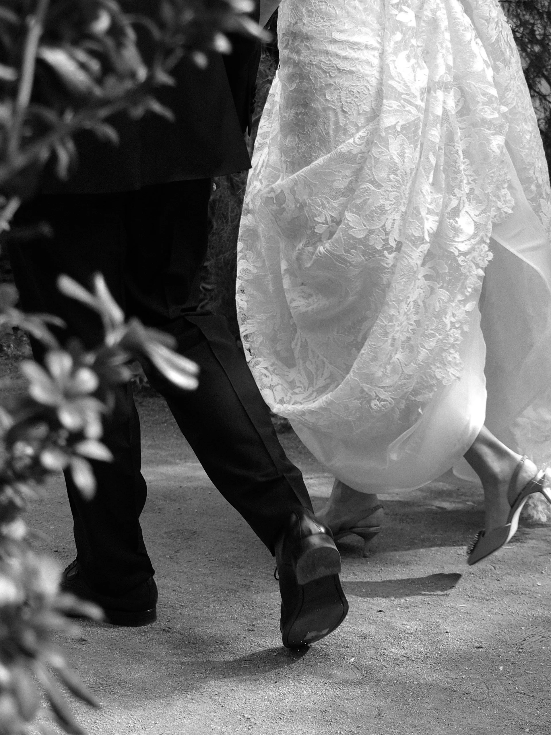 Close-up of a bride and groom dancing, focusing on their feet and lower legs, with the bride wearing an intricate lace wedding dress and high heels, and the groom in dark shoes and pants.
