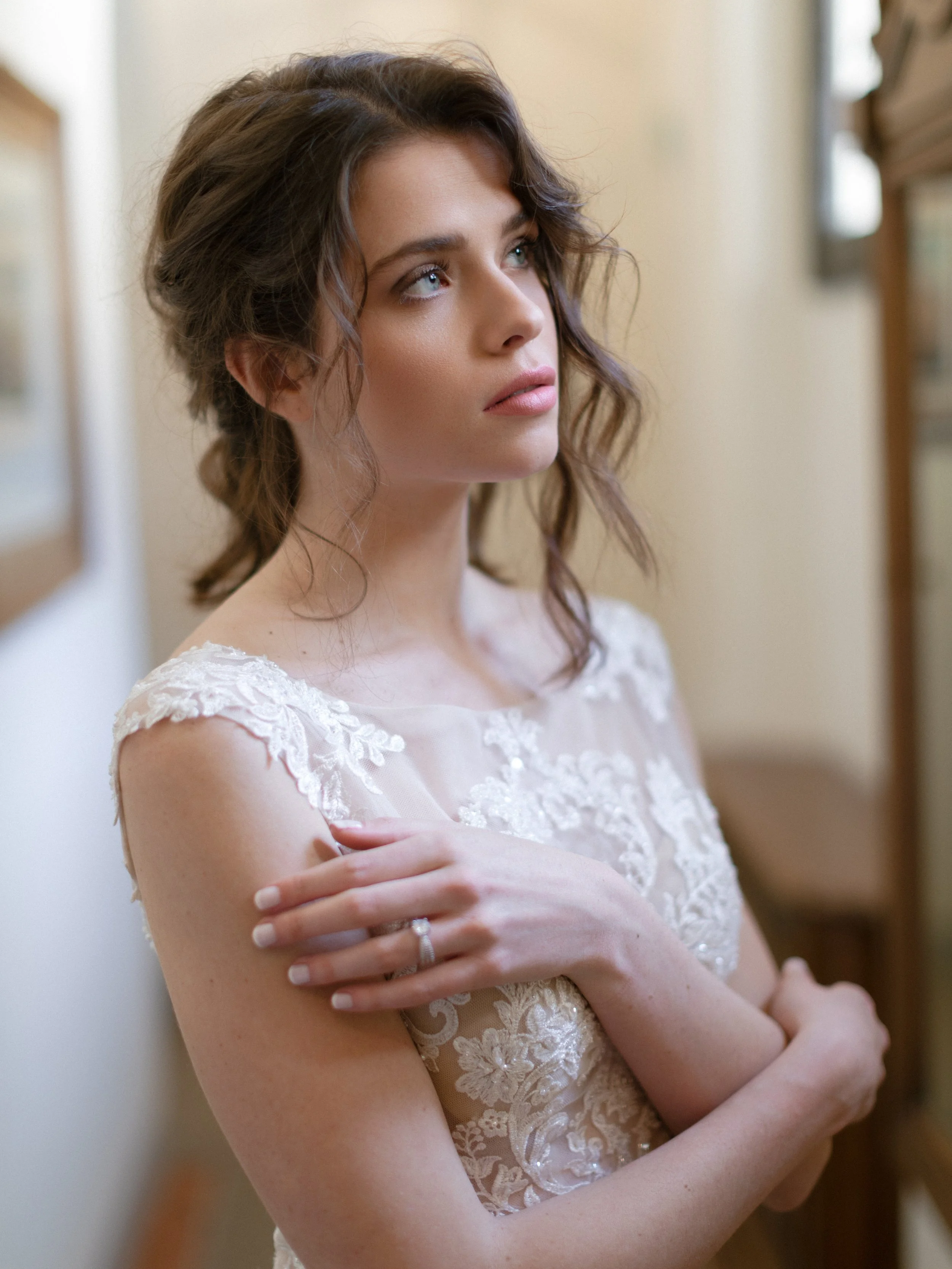 A young woman with wavy brown hair looking thoughtfully to her left, standing indoors near a bookshelf, wearing a white lace dress with floral embroidery.
