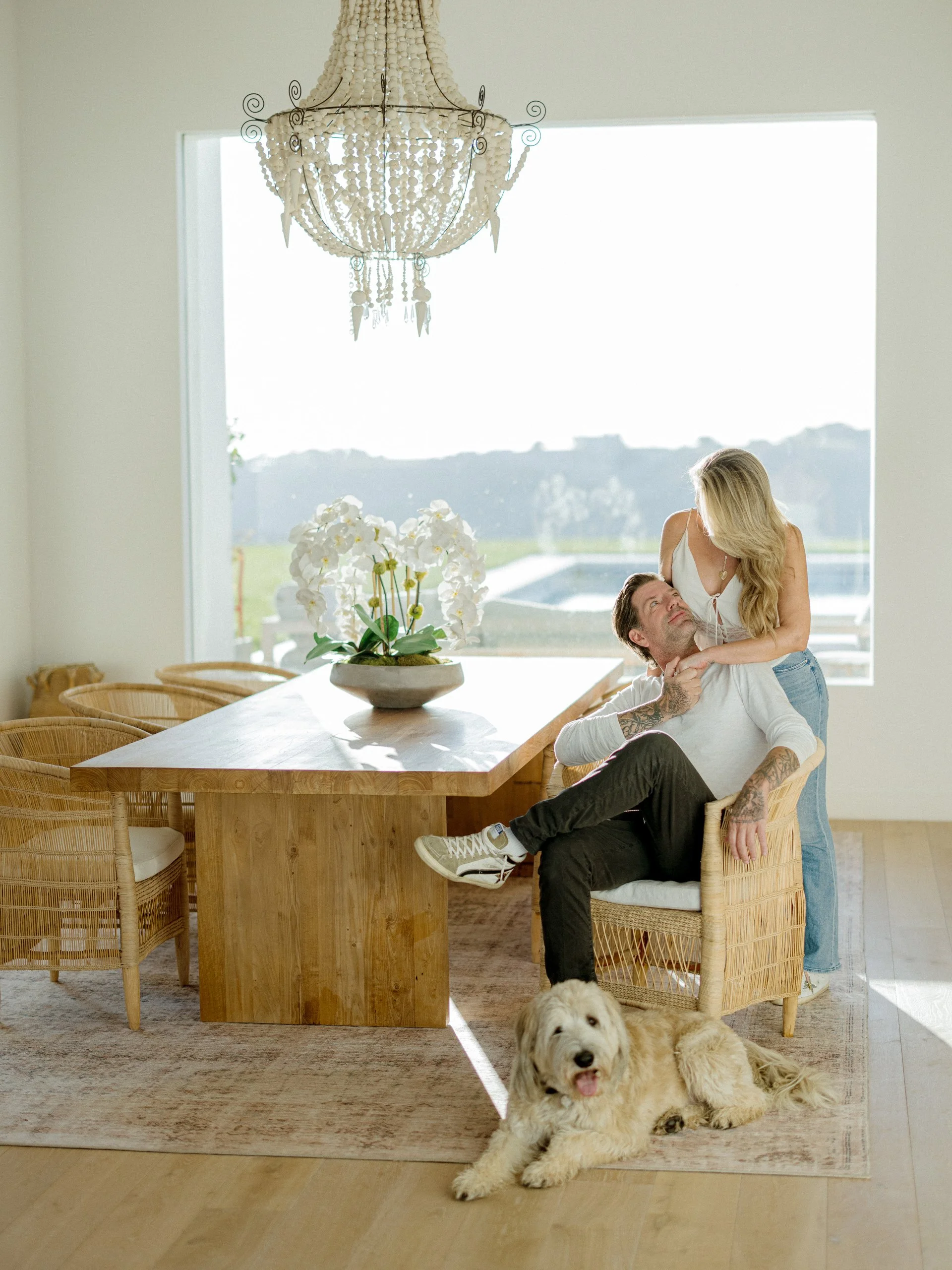 A man sitting in a wicker chair and a woman leaning over him in a dining room with a large window, a chandelier, and a table with a floral centerpiece. There is a dog lying on the floor in front of them.