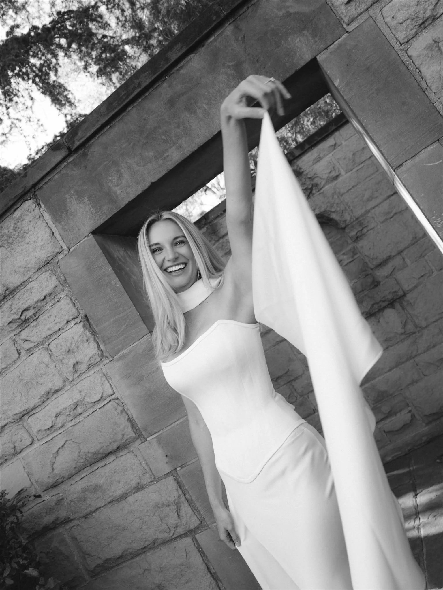 Black and white photo of a smiling woman in a strapless dress holding a flowing piece of fabric, standing outdoors against a stone wall with greenery above.