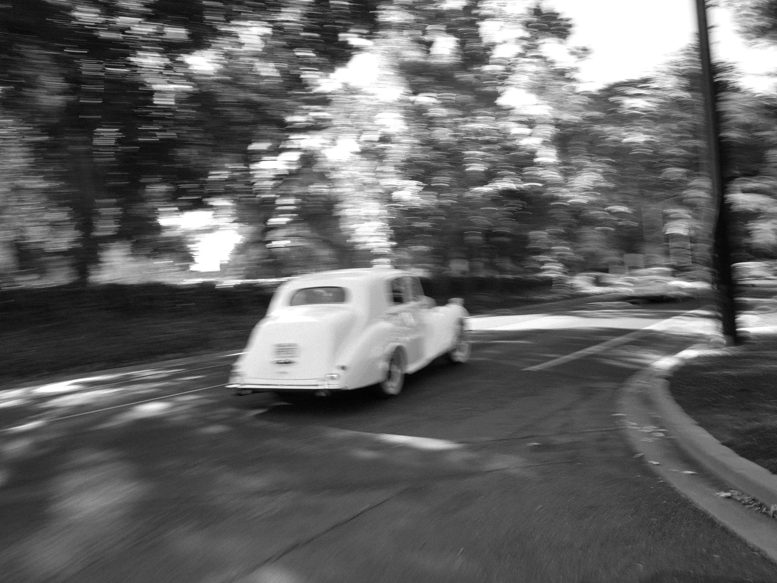 Black and white photo of a vintage car driving on a curved road surrounded by trees.